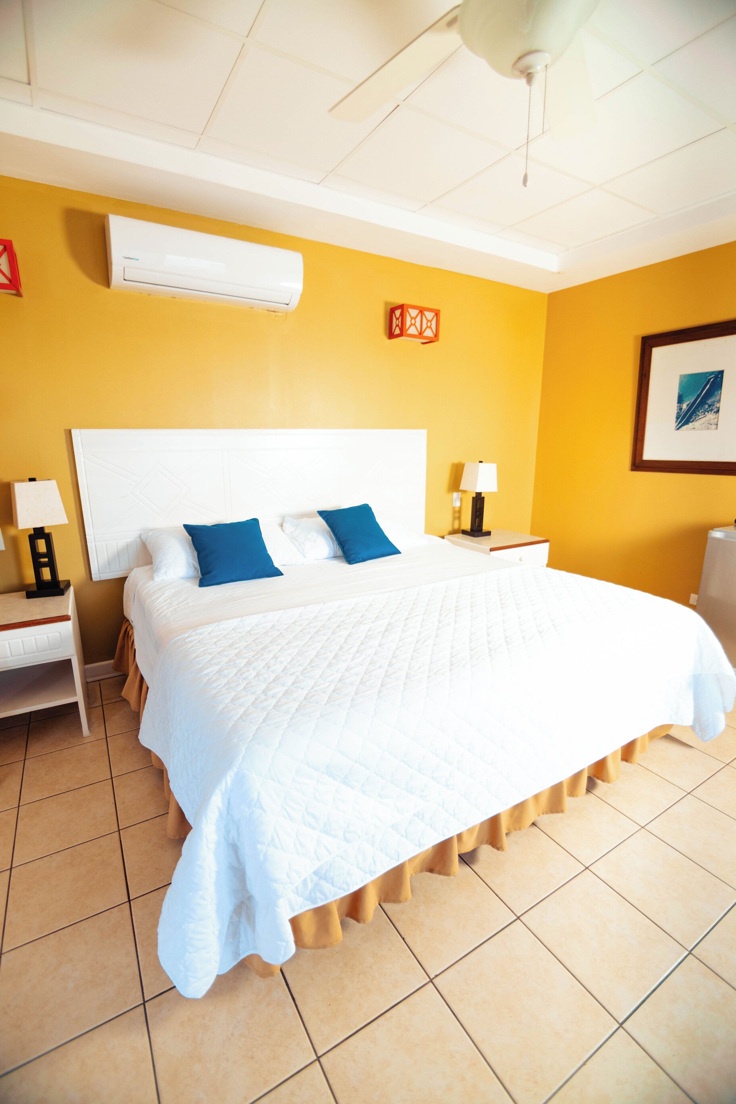 Una habitación de hotel con una cama grande, una mesita de noche, una lámpara, aire acondicionado y ventilador de techo.