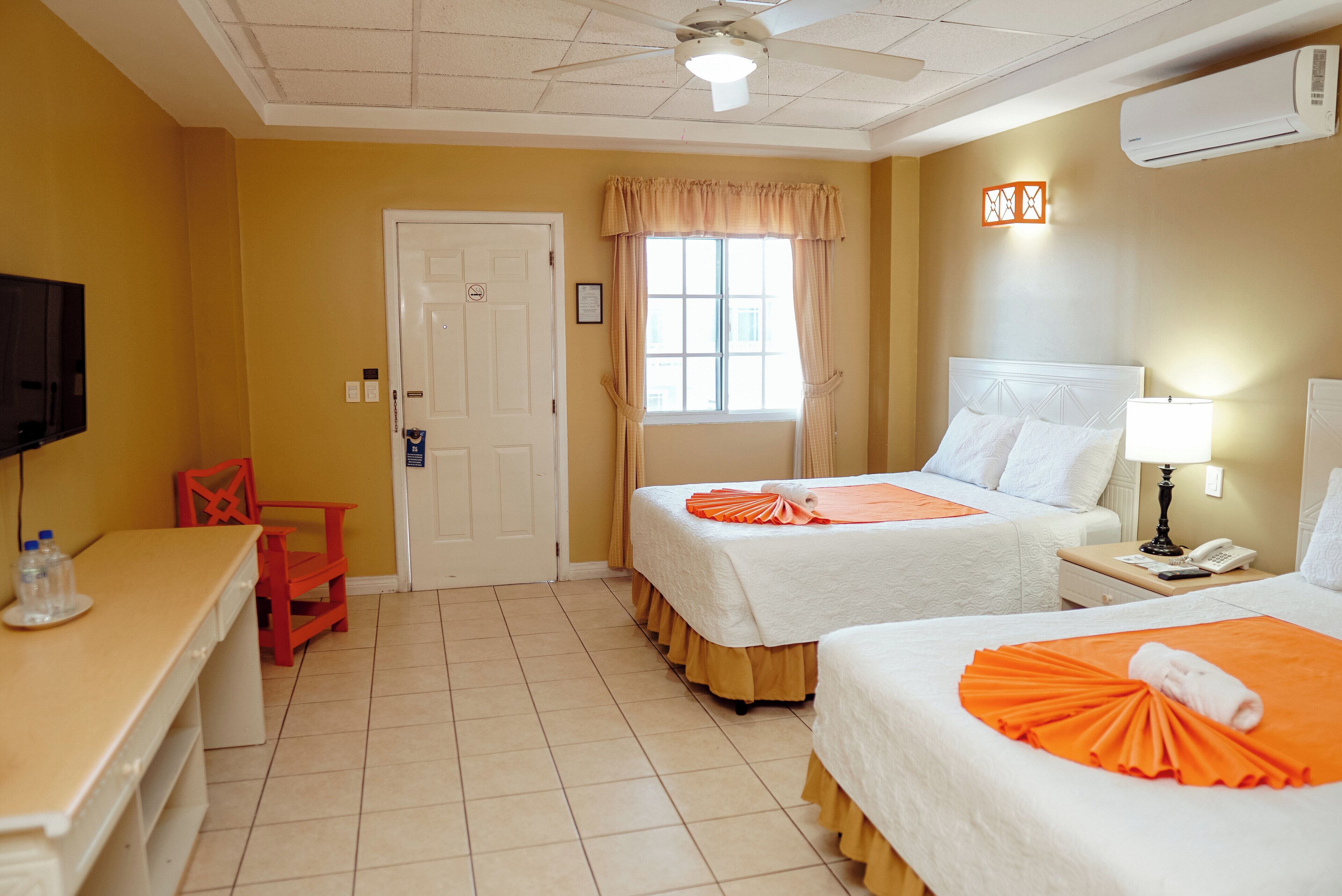 Une chambre d’hôtel avec deux lits, un bureau, une chaise, une fenêtre avec des rideaux, un ventilateur de plafond et un appareil de climatisation.