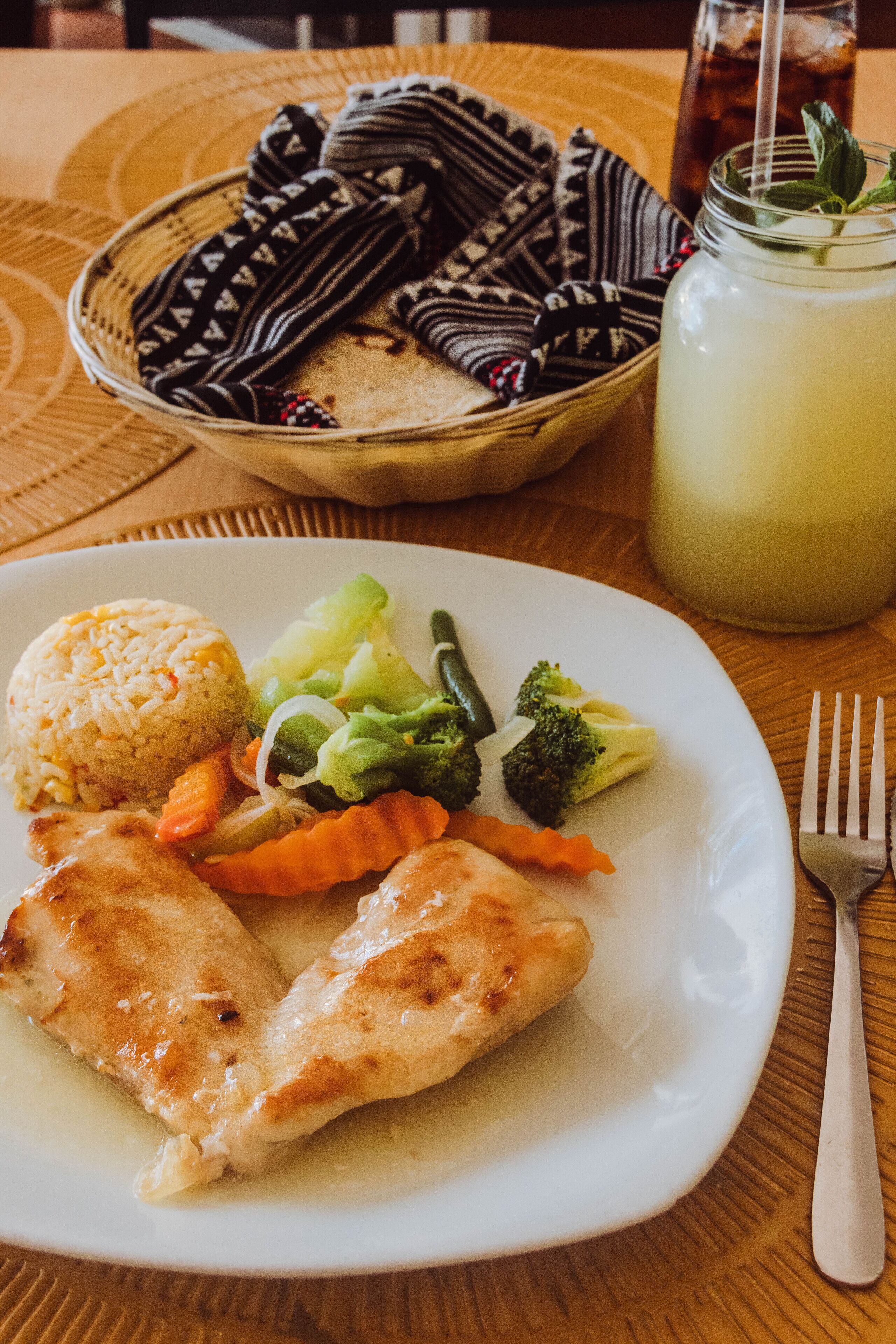 Un plato de comida con arroz, verduras y pollo, un vaso de té helado y una cesta con un paño.