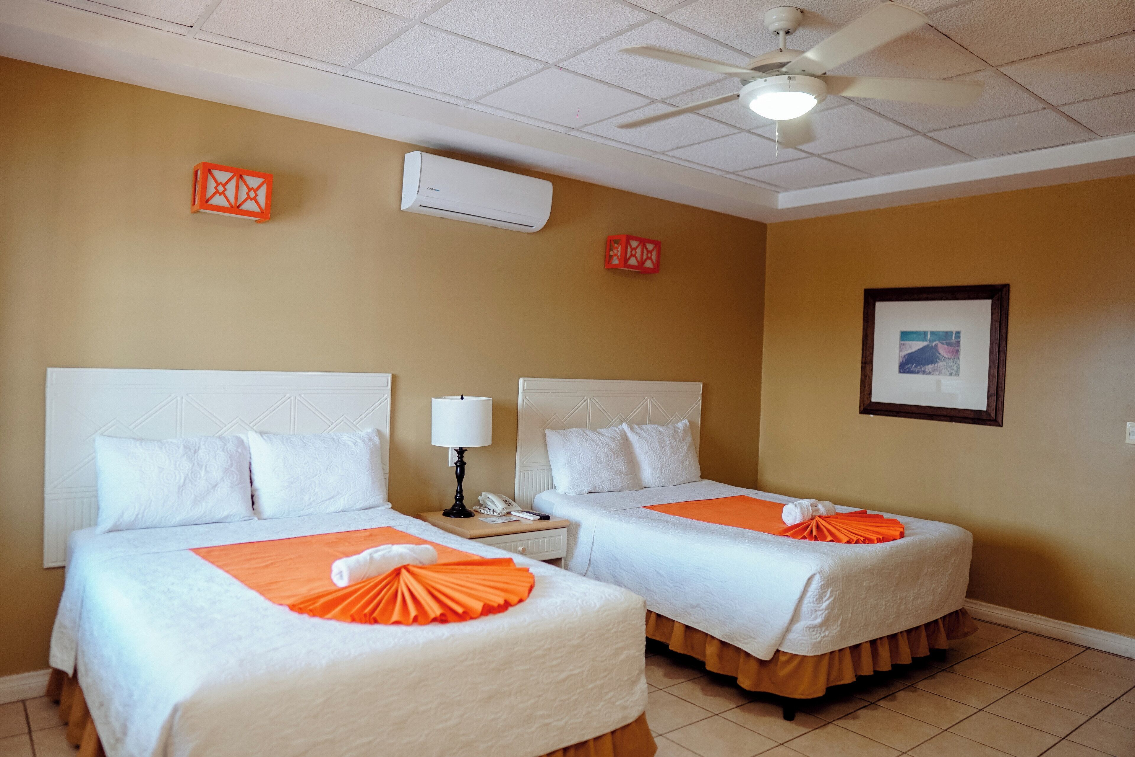 Een hotelkamer met twee bedden, een plafondventilator, een airconditioner, een ingelijst schilderij aan de muur en een telefoon op het nachtkastje.