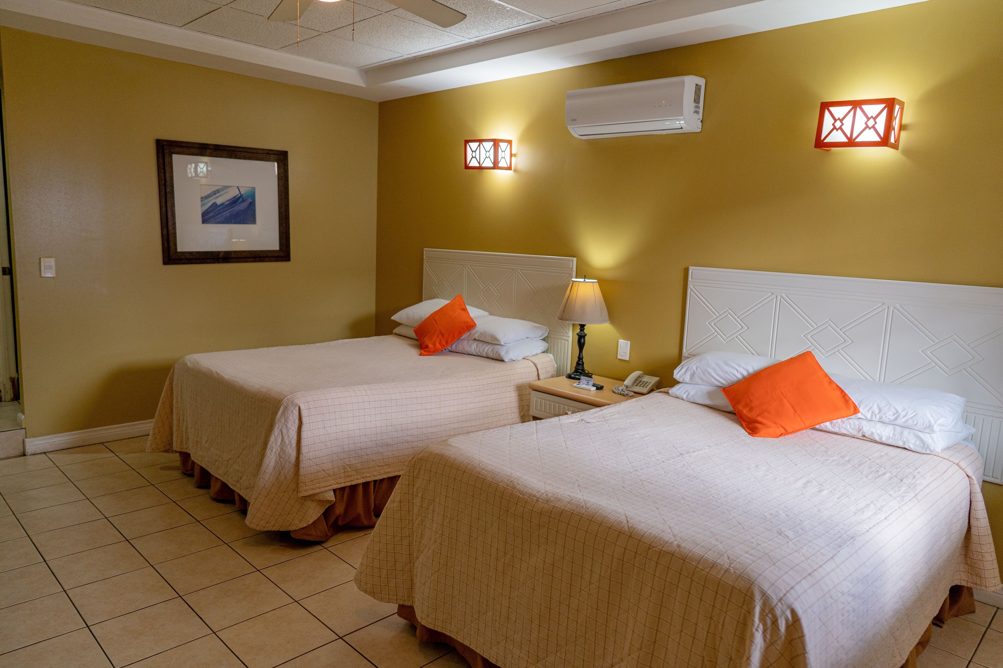 Een hotelkamer met twee bedden, een ingelijst schilderij aan de muur, een airconditioner en een nachtkastje met lamp en telefoon.