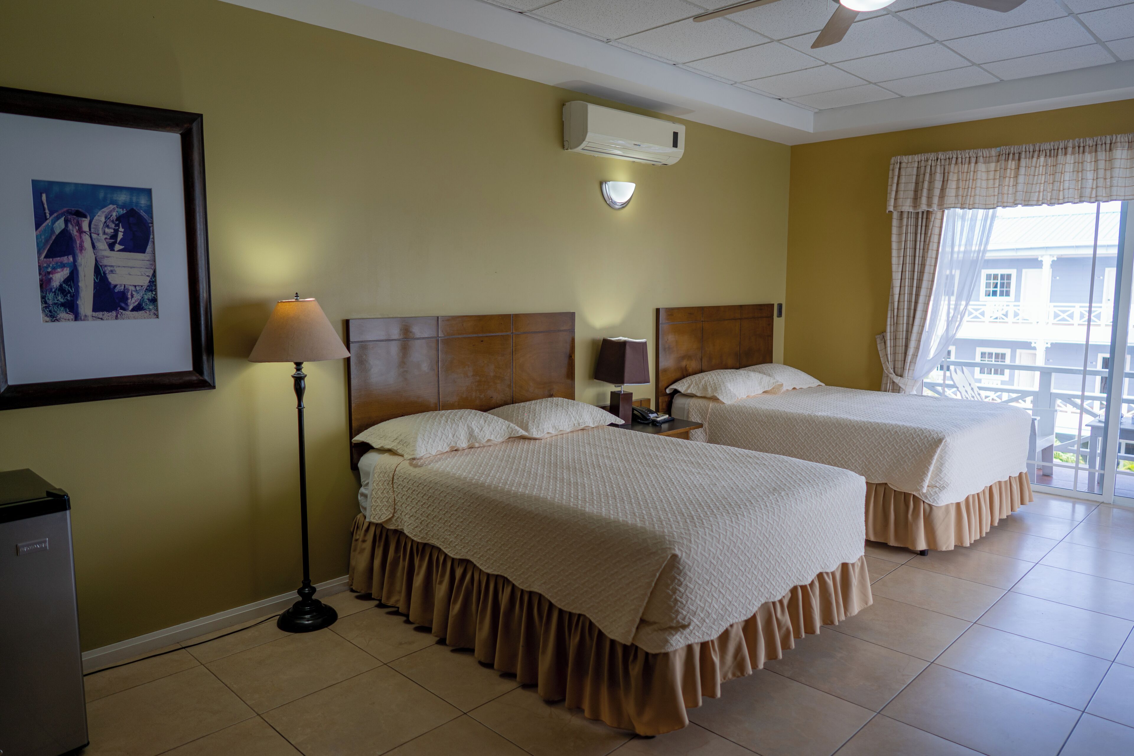 Habitación de hotel con dos camas, ventilador de techo, aire acondicionado, un cuadro enmarcado, una lámpara de pie y ventana con vistas.