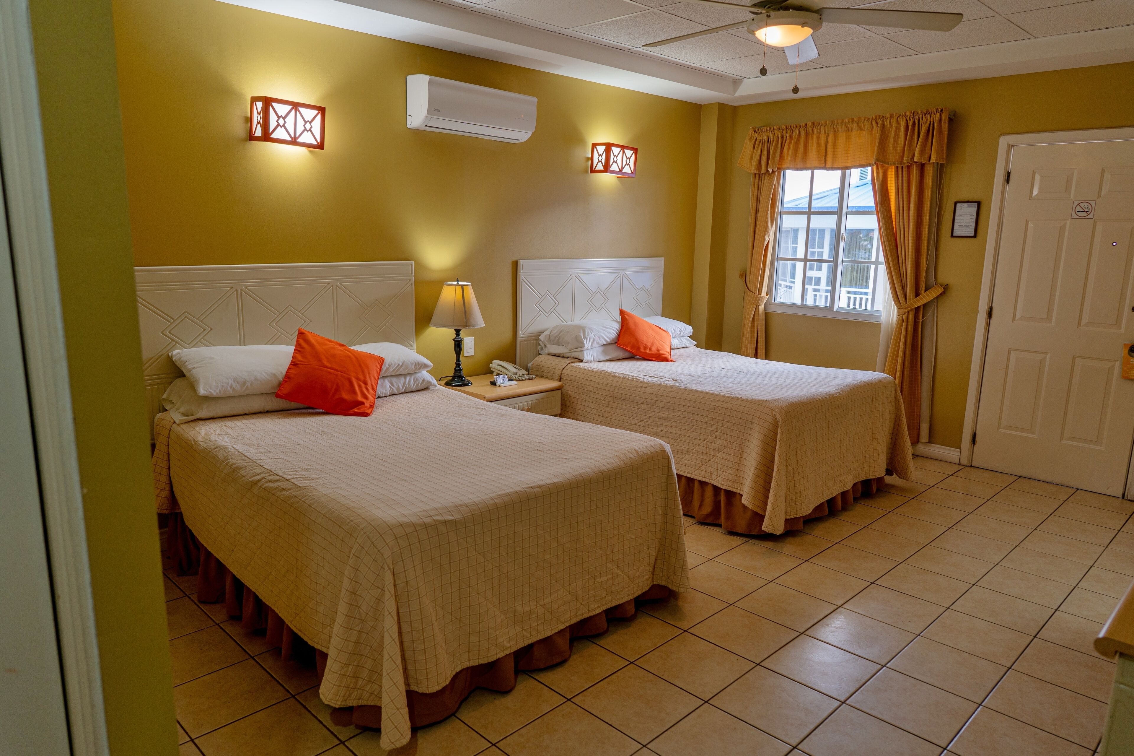 A hotel room with two beds, a ceiling fan, an air conditioning unit, and a window with curtains.