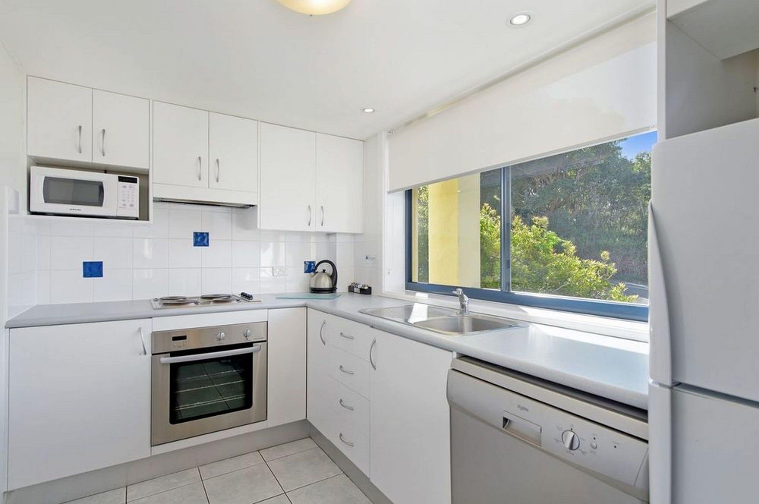 A modern kitchen with white cabinets, a built-in microwave, an oven, a dishwasher, and a large window offering a view of greenery outside.