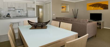 A modern living room with a dining area, a sofa, a flat-screen TV, and a kitchen in the background.