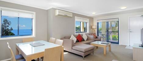 A modern living room with a sofa, coffee table, and dining table. There are large windows with a view of the sea.