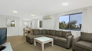 A modern living room with a sectional sofa, a glass coffee table, and a TV on a stand. There is a dining area with a table and chairs, and large windows with curtains.