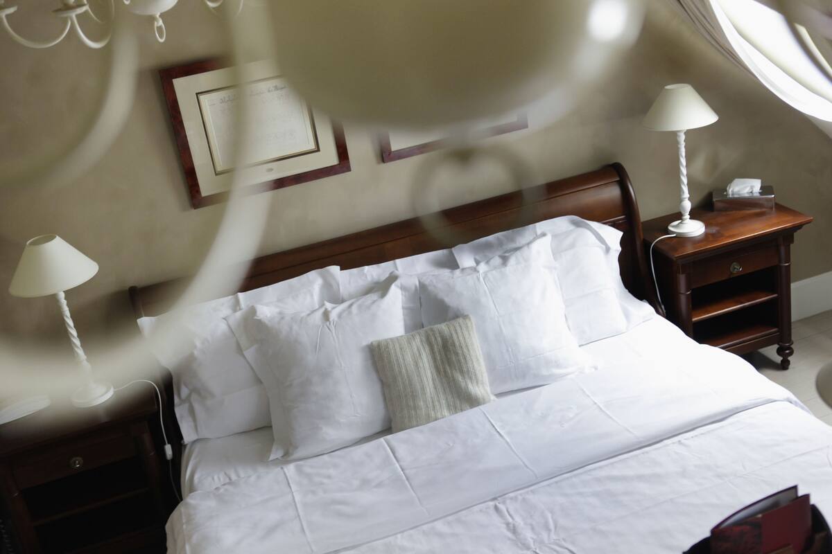 Un lit bien fait, avec du linge de lit blanc et des oreillers, une tête de lit en bois, une table de chevet avec une lampe et un certificat encadré accroché au mur.