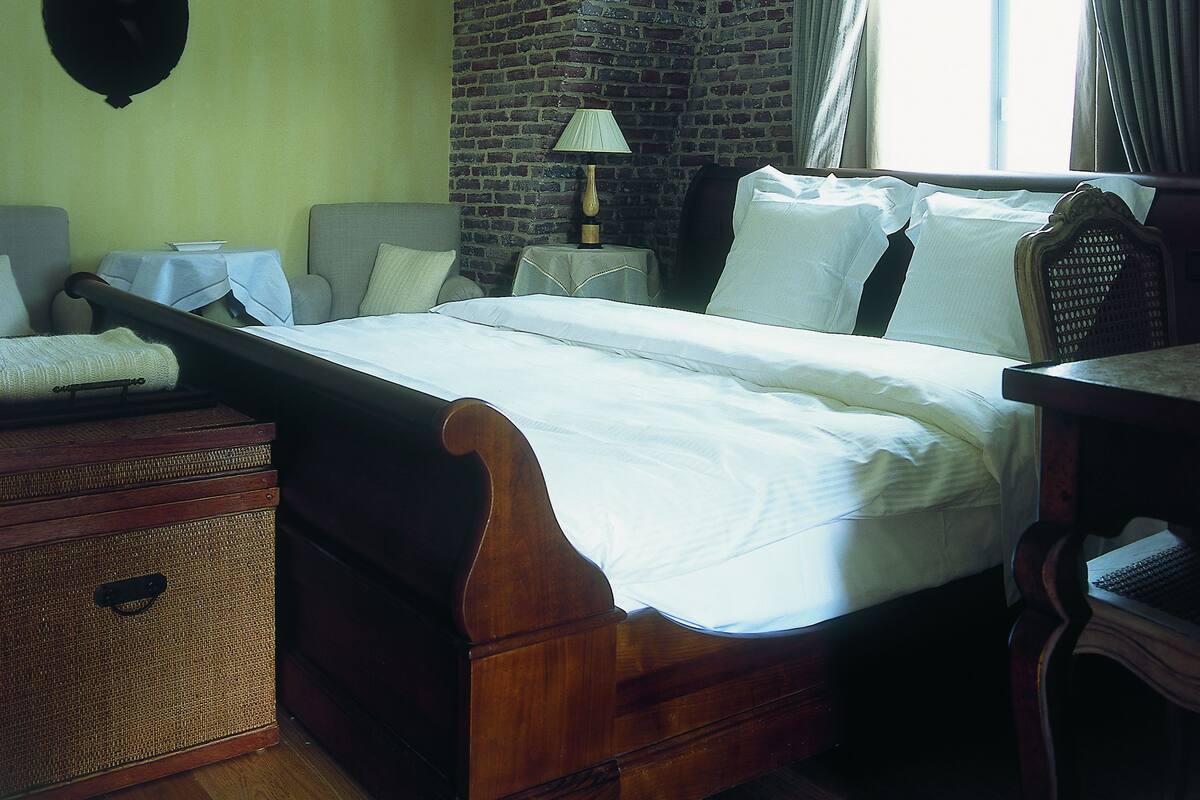 Une chambre à l’ancienne avec un mur en briques, un lit recouvert de draps blancs, une tête de lit en bois, une table de chevet avec une lampe et une fenêtre avec des rideaux.