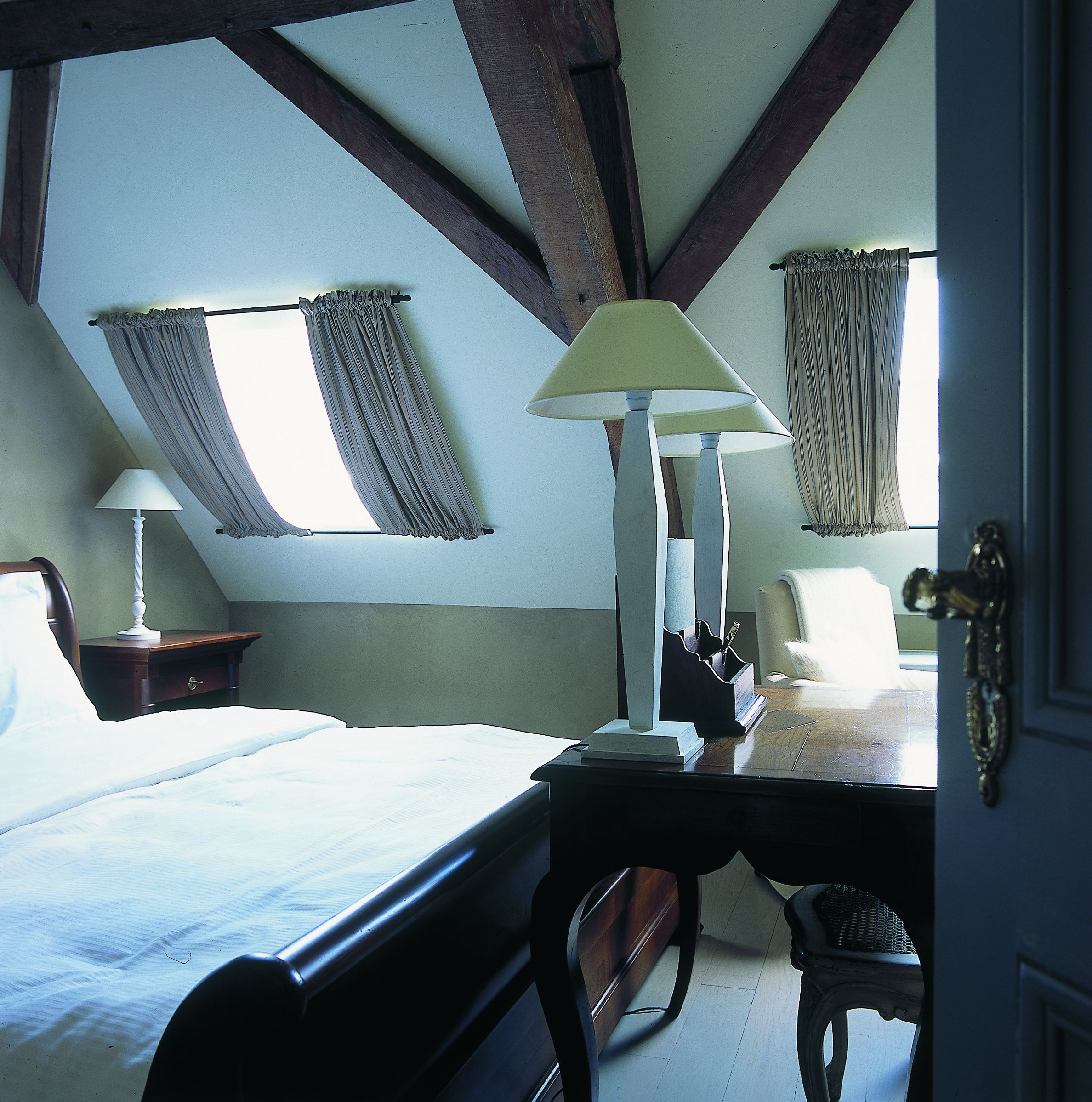 Ein Schlafzimmer mit einem Bett, einem Schreibtisch, einer Lampe und zwei Fenstern mit Vorhängen.