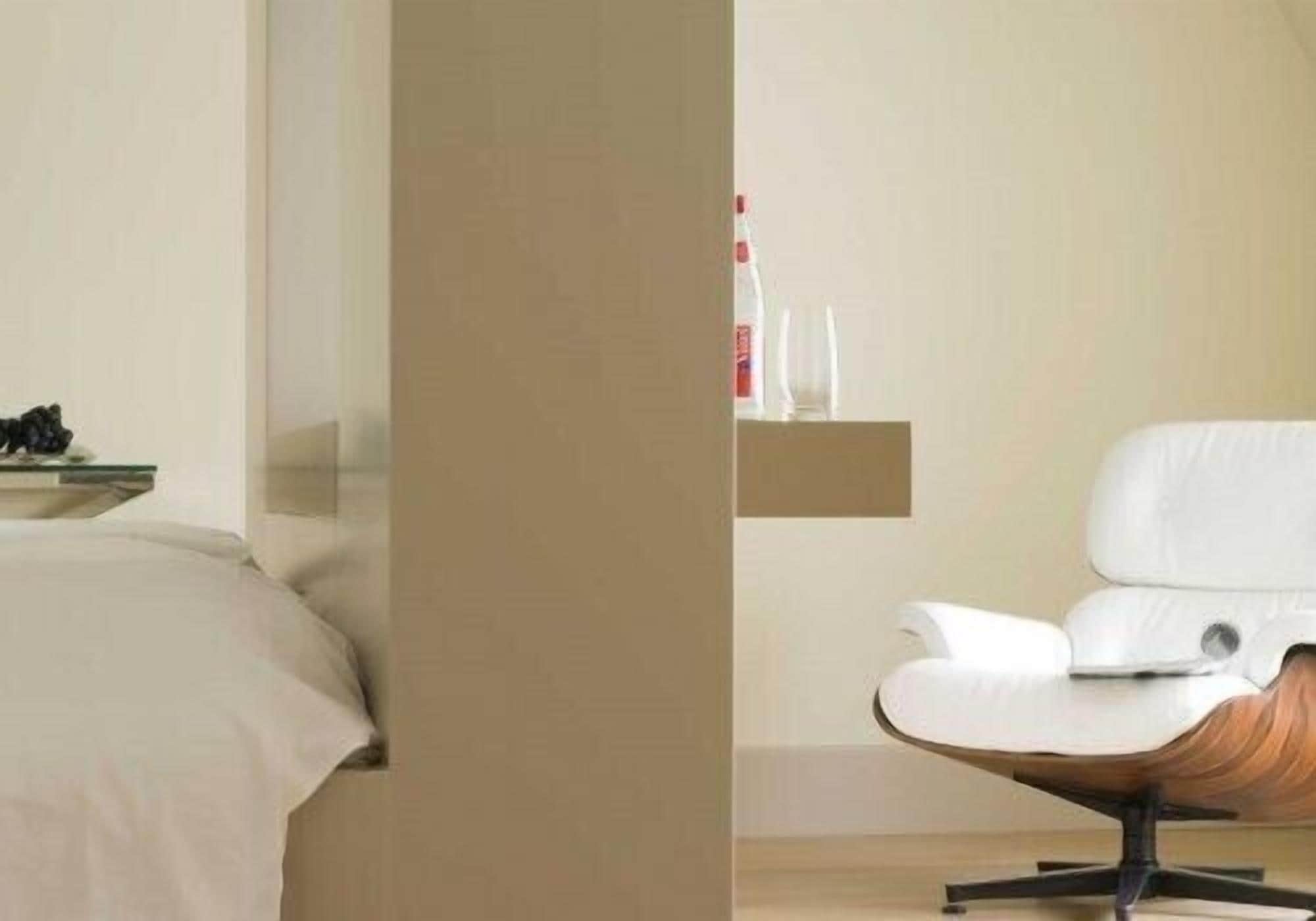 Une chambre d’hôtel moderne avec un lit, une chaise blanche et une petite étagère sur laquelle se trouvent une bouteille et un verre.