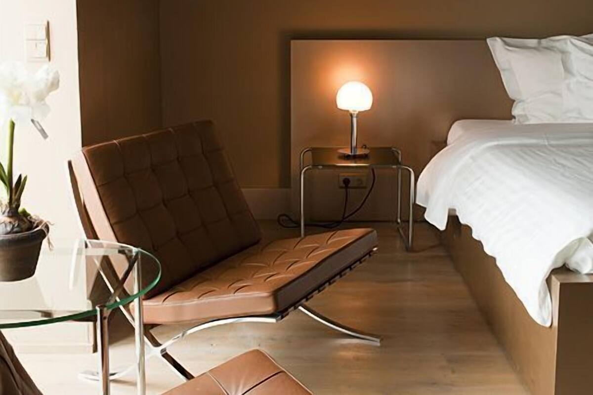 Une chambre à coucher avec un lit, une table de chevet avec une lampe, un fauteuil rembourré et une plante en pot.
