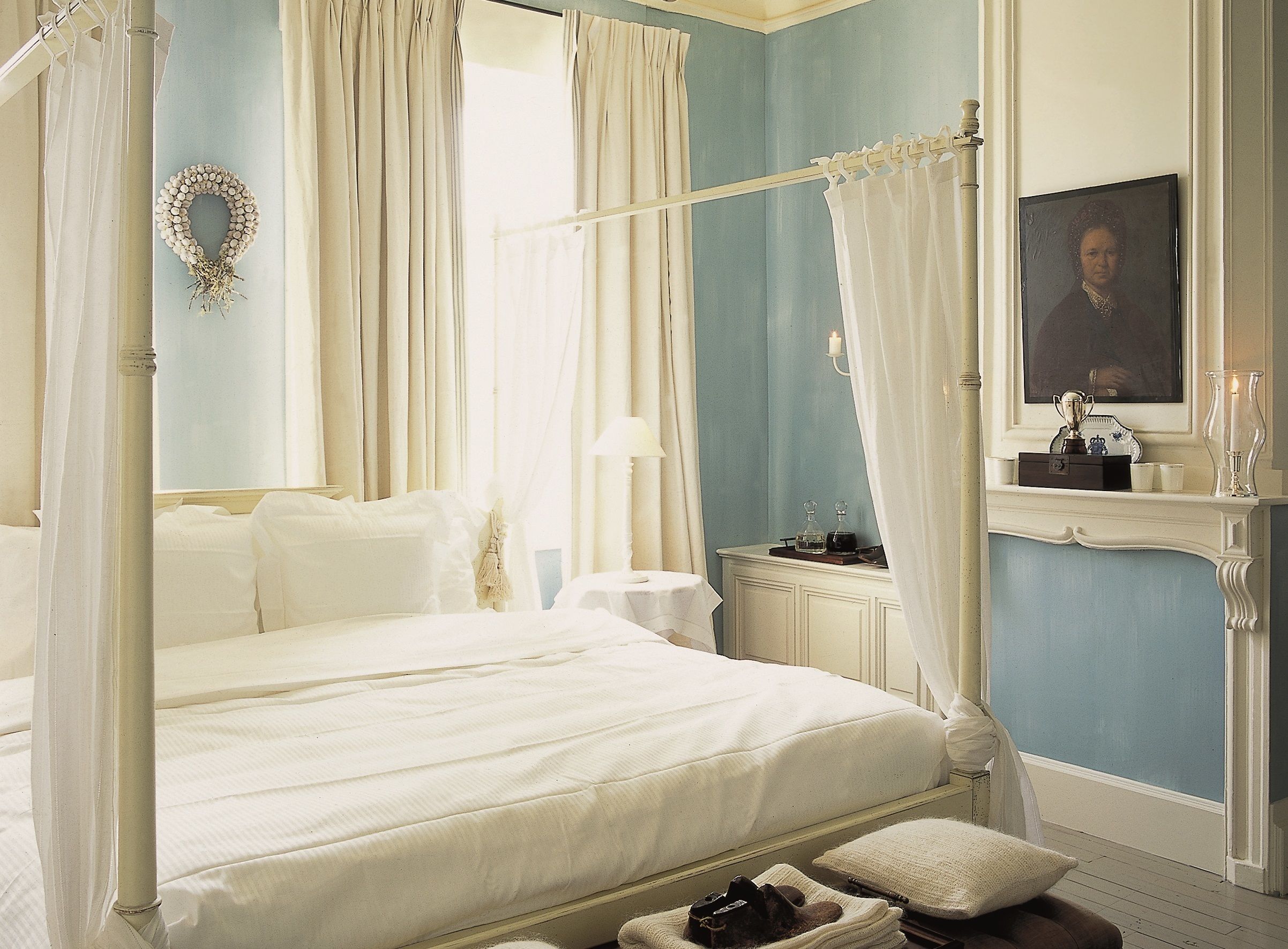 A four-poster canopy bed with white linens, a bedside table with a lamp, and a mirror above a fireplace with a framed portrait and a vase.