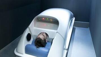 A medical treatment room with a reclining treatment bed, a control panel, and a ceiling light.