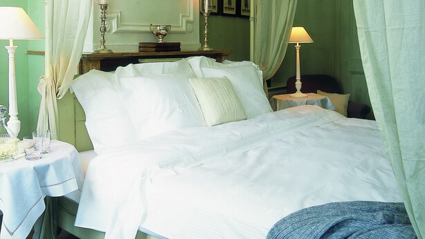 A four-poster bed with white linens, a bedside table with a lamp, and a mirror above the bed.