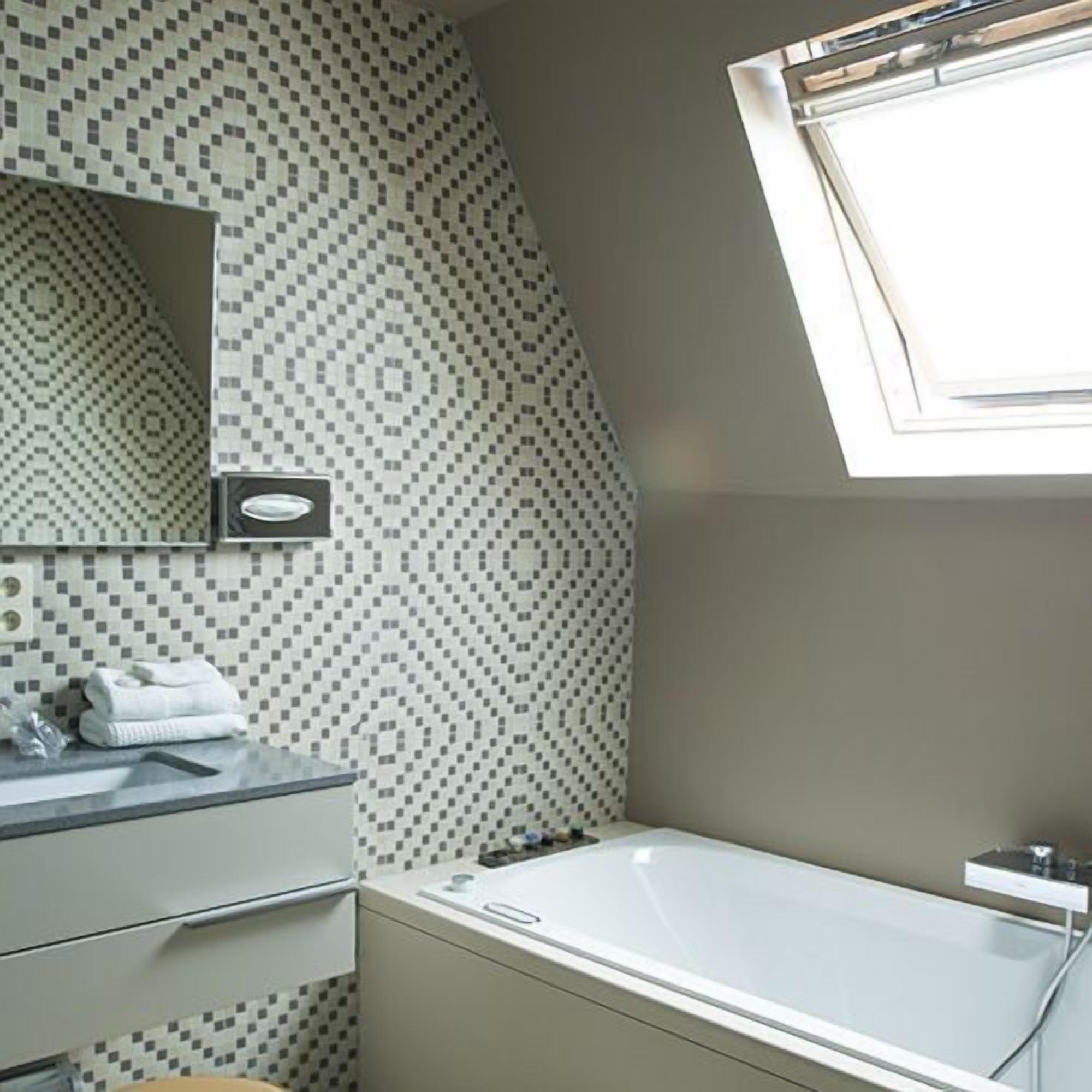 A modern bathroom with a geometric patterned wall, a built-in sink, a mirror, and a window above the bathtub.