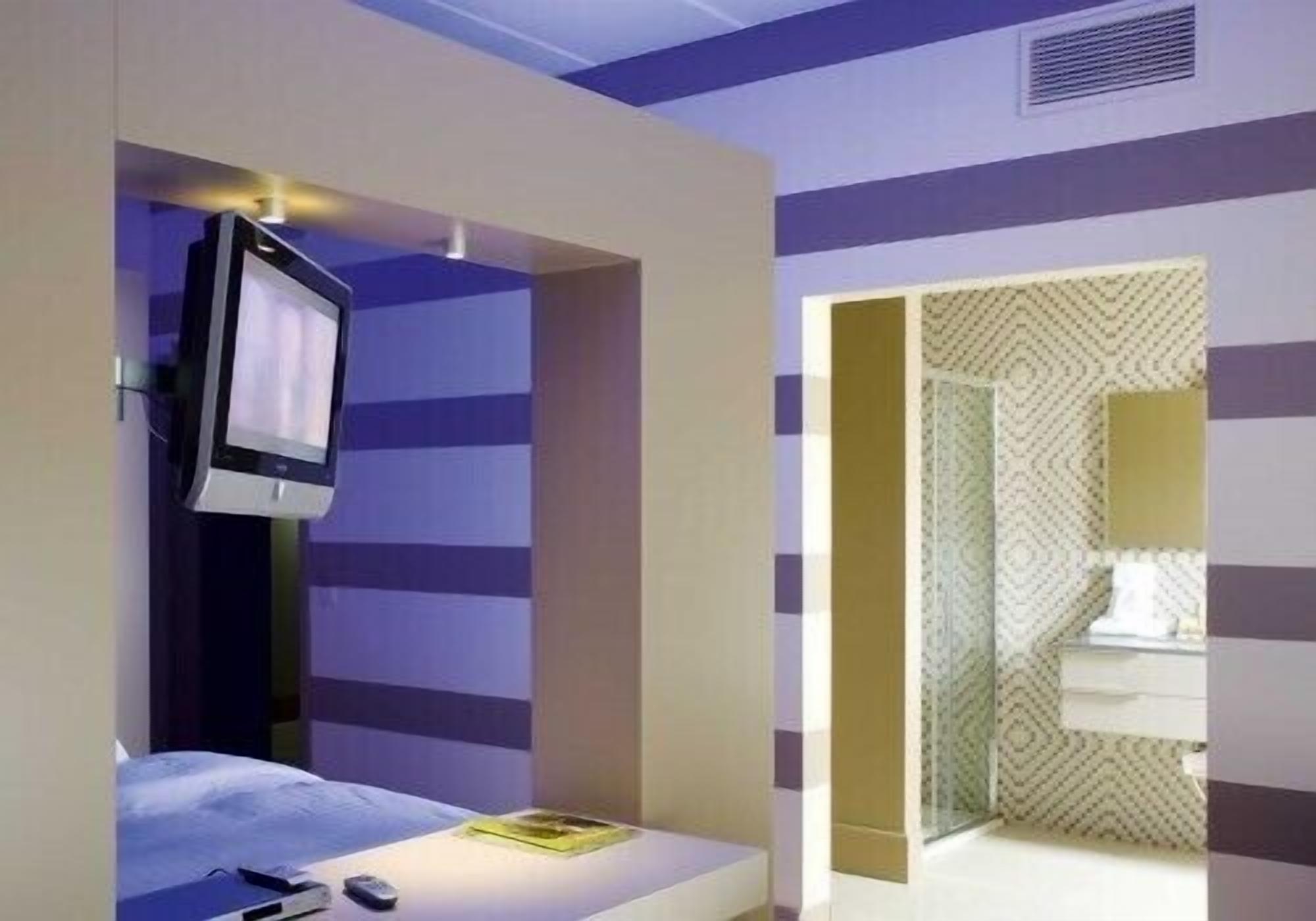 A hotel room with a bed, a TV mounted on the wall, and a bathroom with patterned wallpaper.