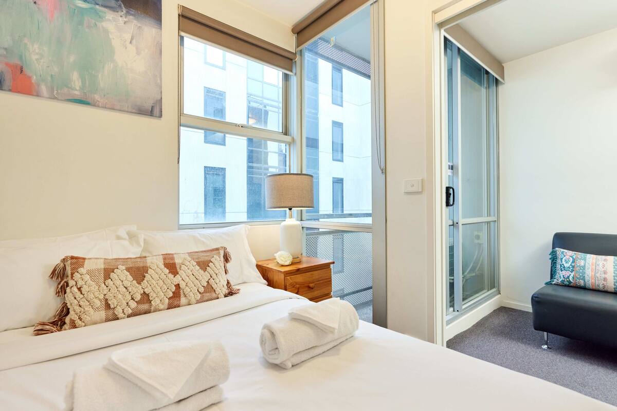 Habitación de hotel con cama, mesita de noche, lámpara, ventana con vistas a edificios y puerta a un balcón.