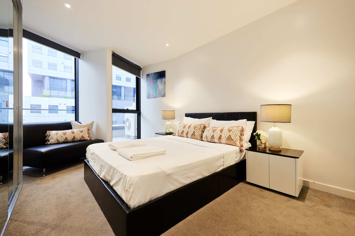 Un dormitorio moderno con una cama grande, una mesita de noche con lámparas, un sofá y un amplio ventanal con vistas a la ciudad.