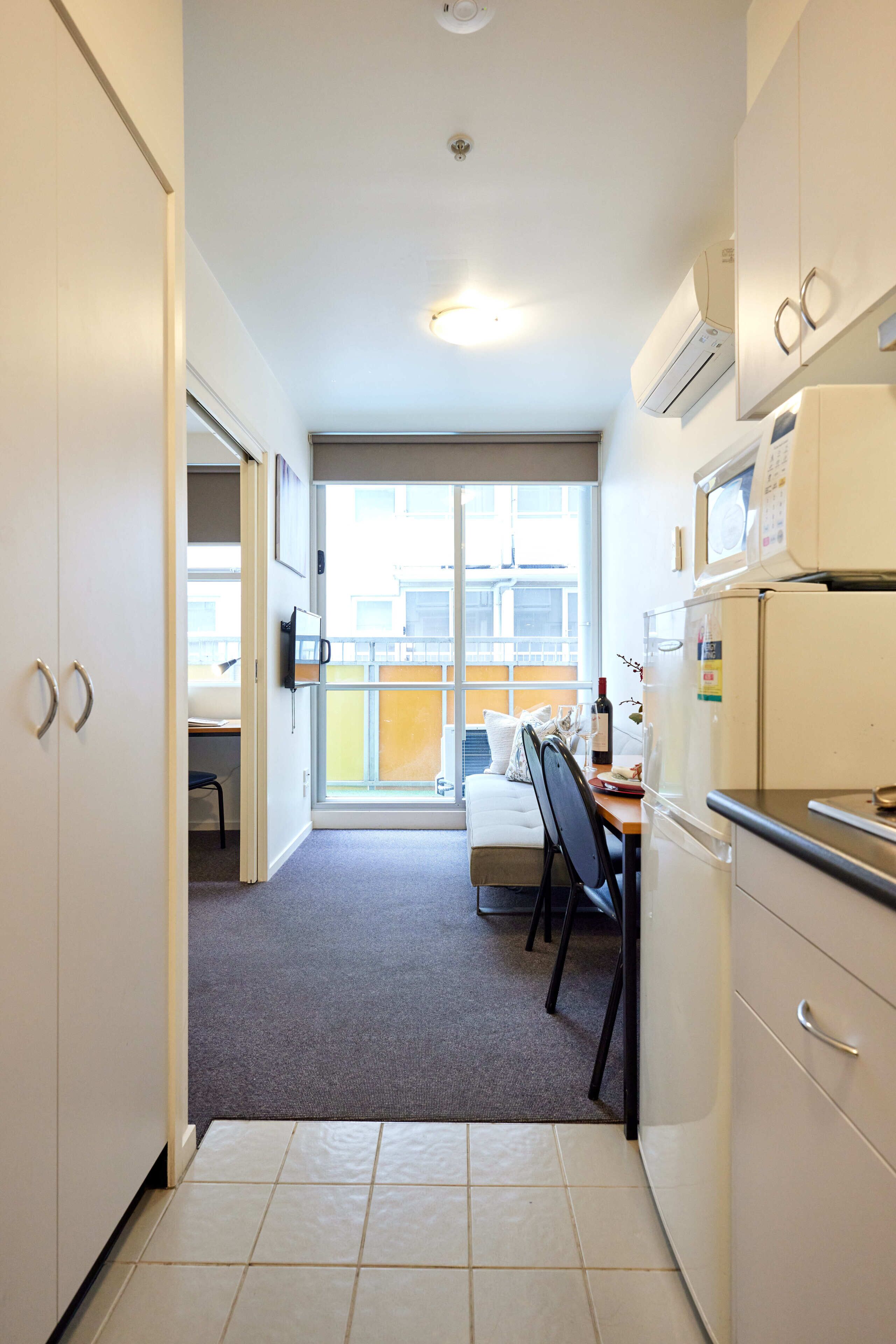 Cocina compacta con armarios blancos, un microondas y un refrigerador. Cuenta con un comedor con mesa y sillas, y una ventana que ofrece vistas al exterior.