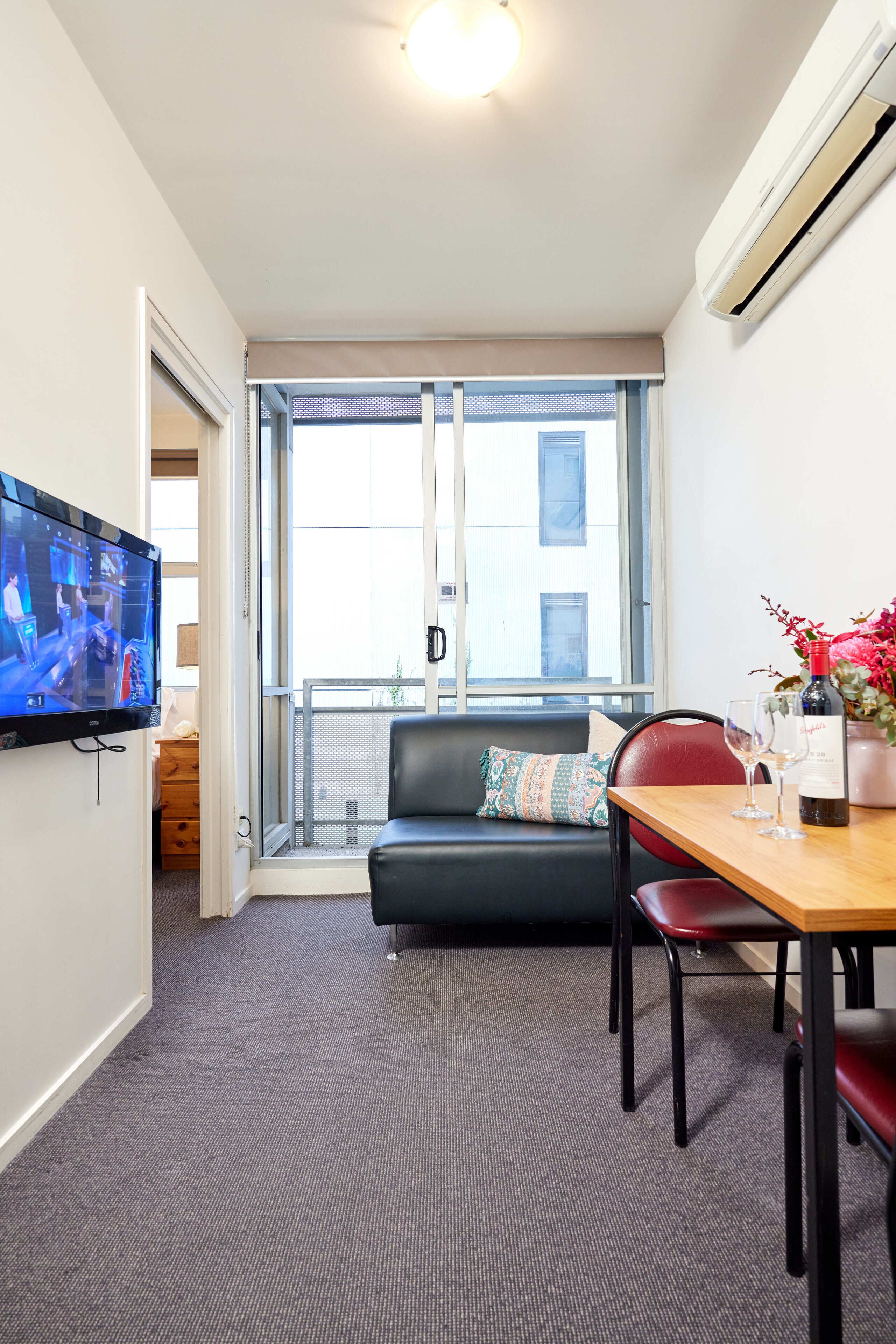 Un salon moderne avec un canapé en cuir noir, une table à manger en bois accompagnée de deux chaises, un téléviseur à écran plat fixé au mur et une grande fenêtre offrant une vue sur la ville.