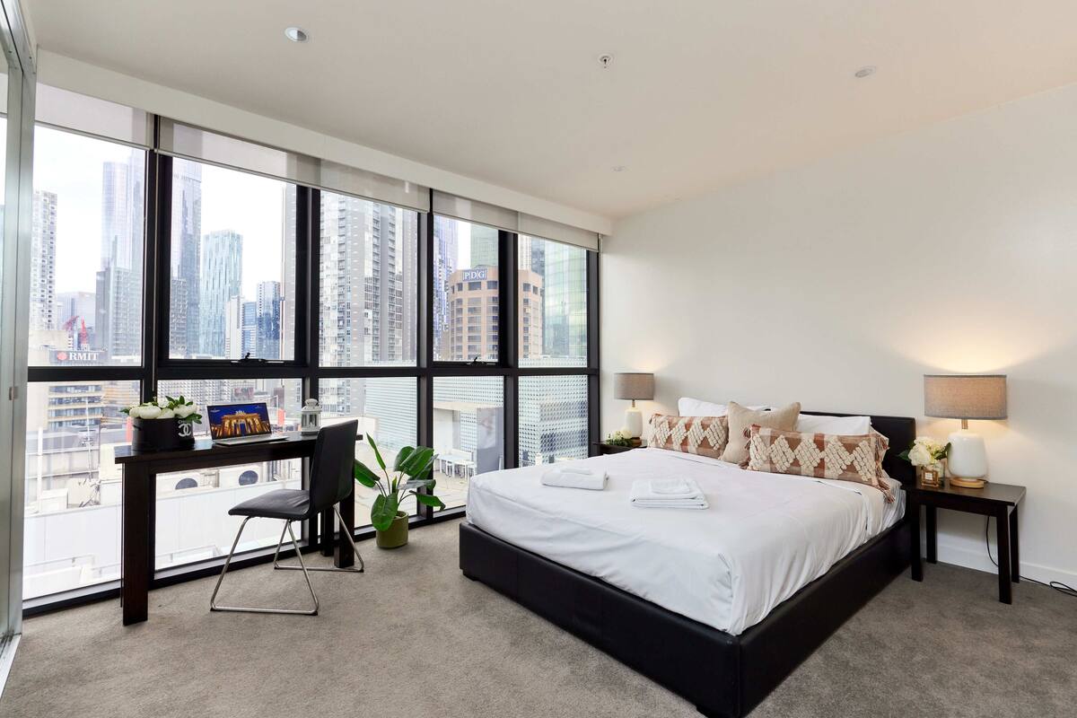 Habitación de hotel moderna con una cama grande, un escritorio y vistas de la silueta urbana a través de ventanales del suelo al techo.