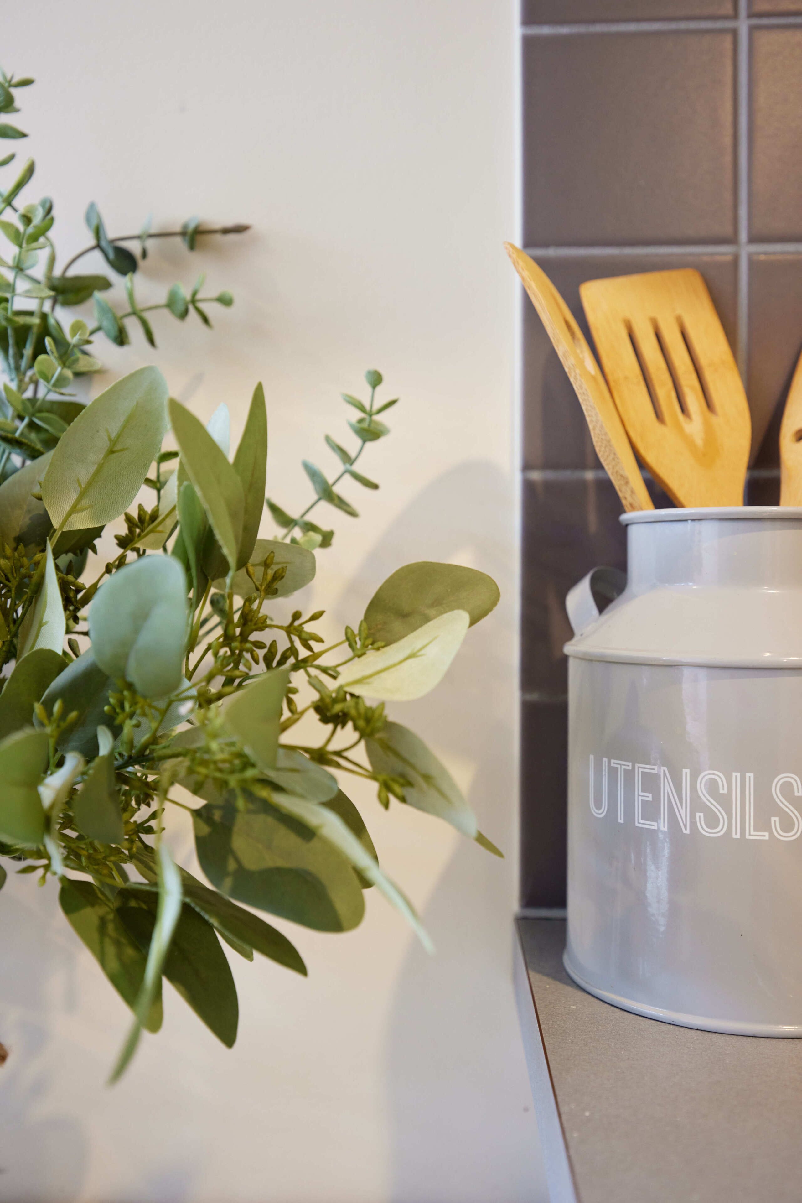 Eine Küchenarbeitsplatte mit einem weißen Utensilienhalter mit der Aufschrift 'UTENSILS' und einem Topf mit Grünpflanzen.