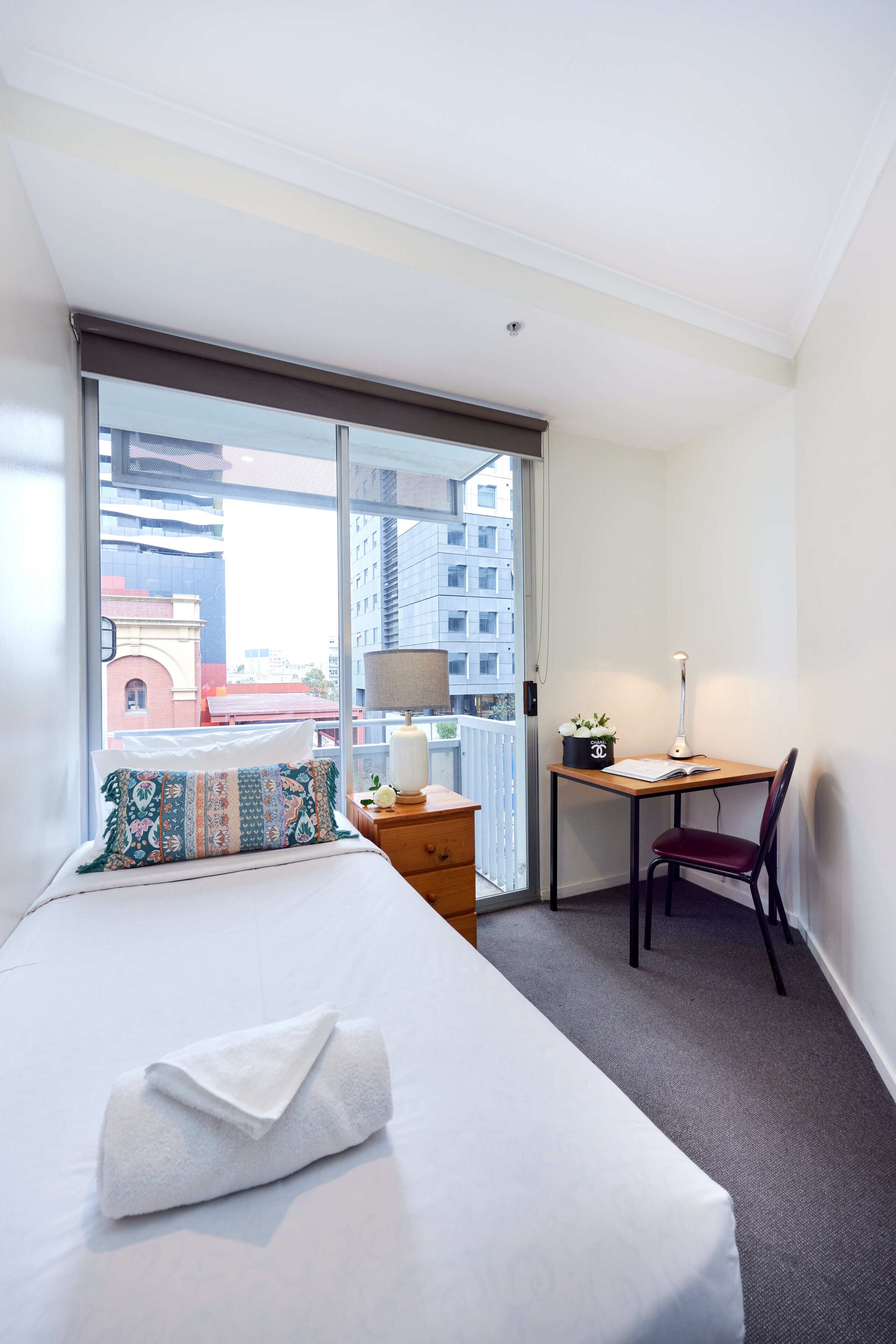 Habitación de hotel con cama, escritorio, silla, lámpara y vista de la ciudad a través de un ventanal.