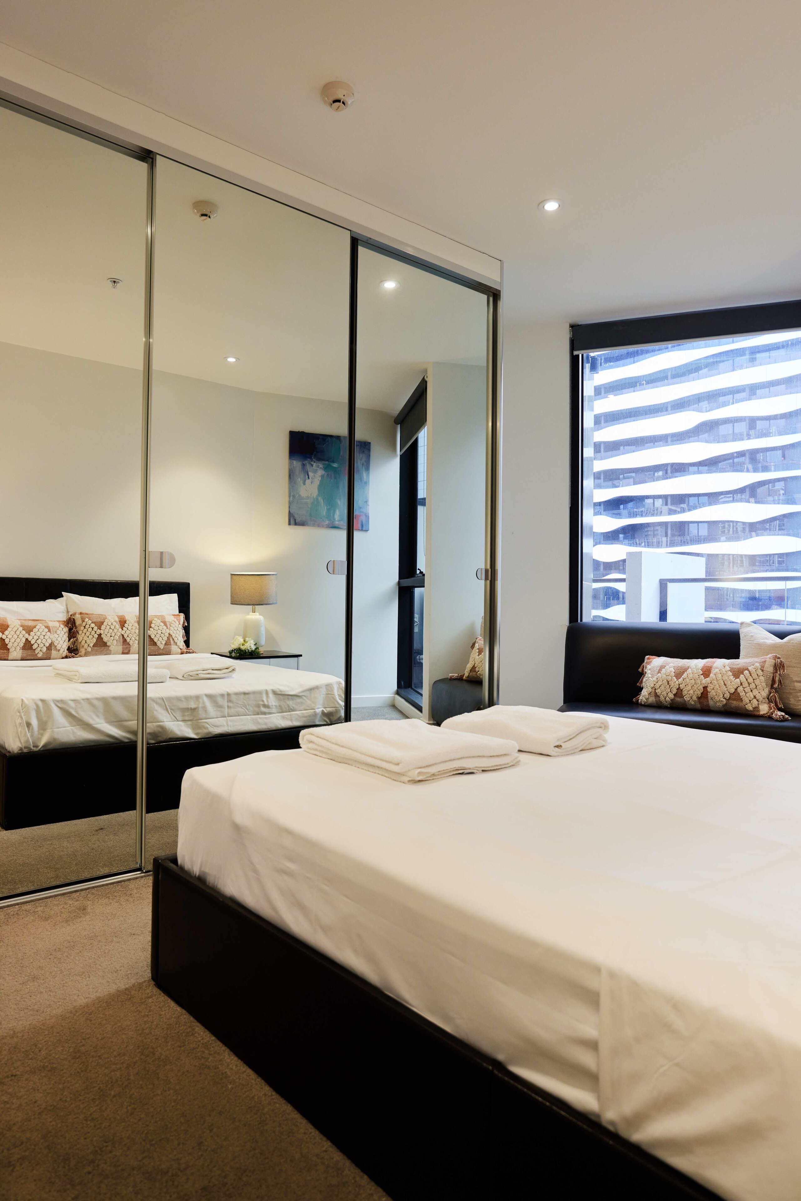 A modern hotel room with a large bed, a glass partition wall, and a view of a building outside.