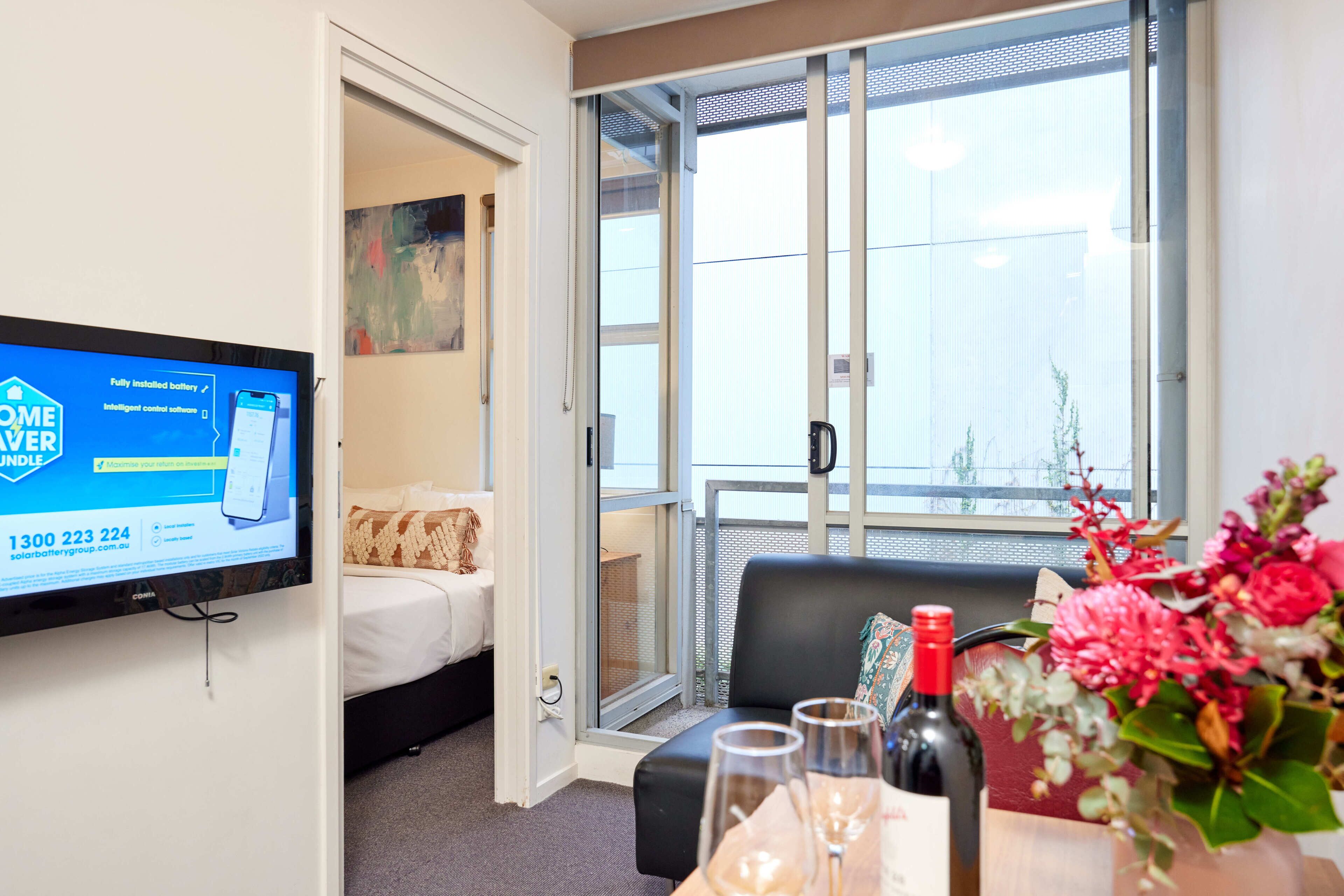 Habitación de hotel con una cama, un televisor mostrando un anuncio de servicio de batería, un balcón con vistas y una mesa con una botella y vasos.