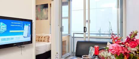 Habitación de hotel con una cama, un televisor mostrando un anuncio de servicio de batería, un balcón con vistas y una mesa con una botella y vasos.