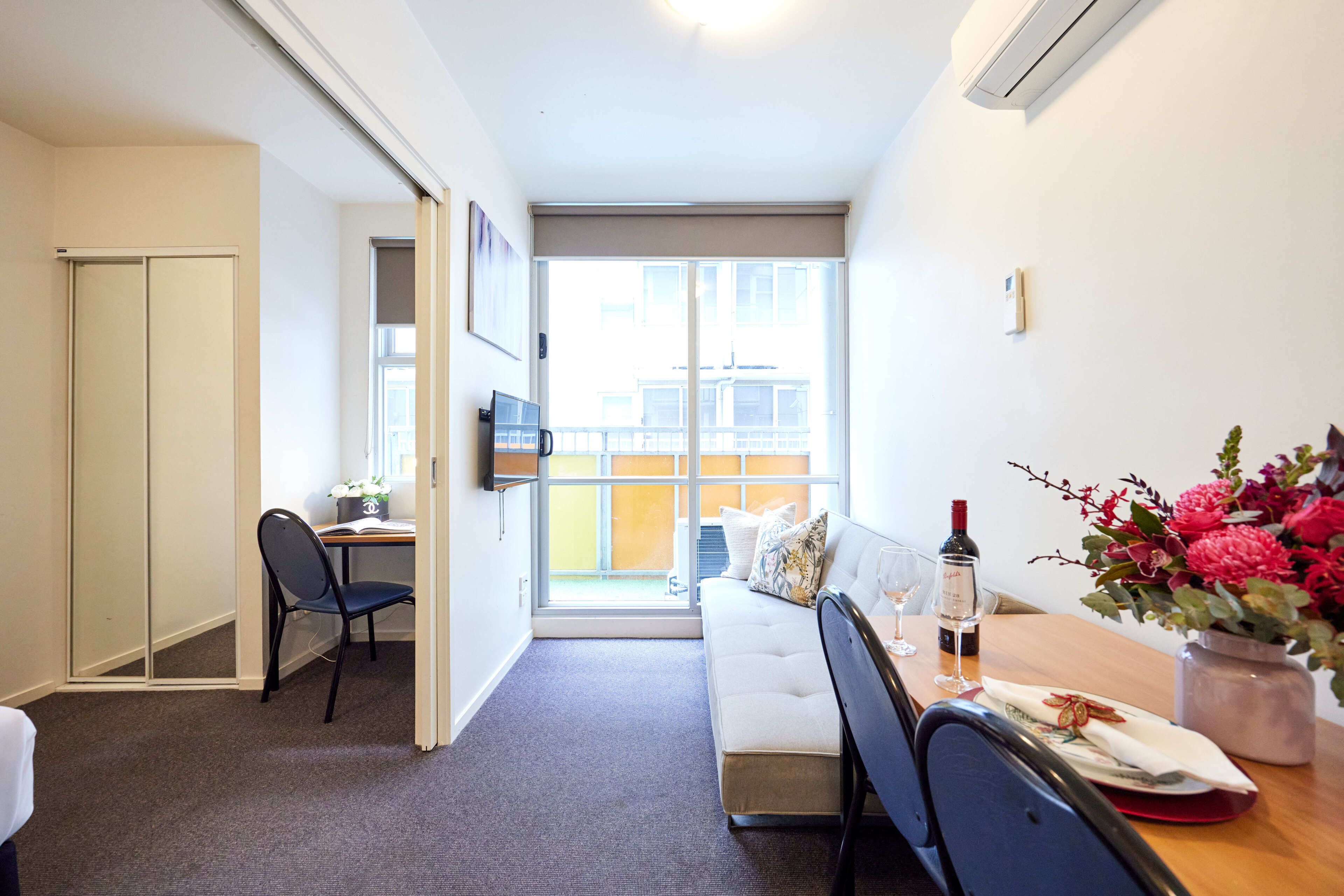 A modern hotel room with a sliding door to a balcony, a sofa, a dining table with a vase of flowers, and a TV on the wall.