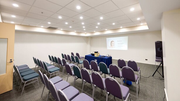 Una sala de conferències amb files de cadires, una pantalla de projector i una taula amb un ordinador portàtil i material de presentació.