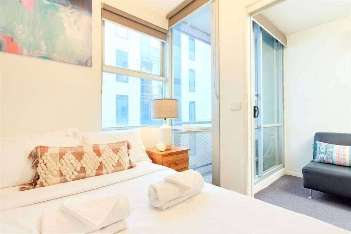 Una habitación de hotel con una cama, una mesita de noche con lámpara, un sofá y un ventanal grande.