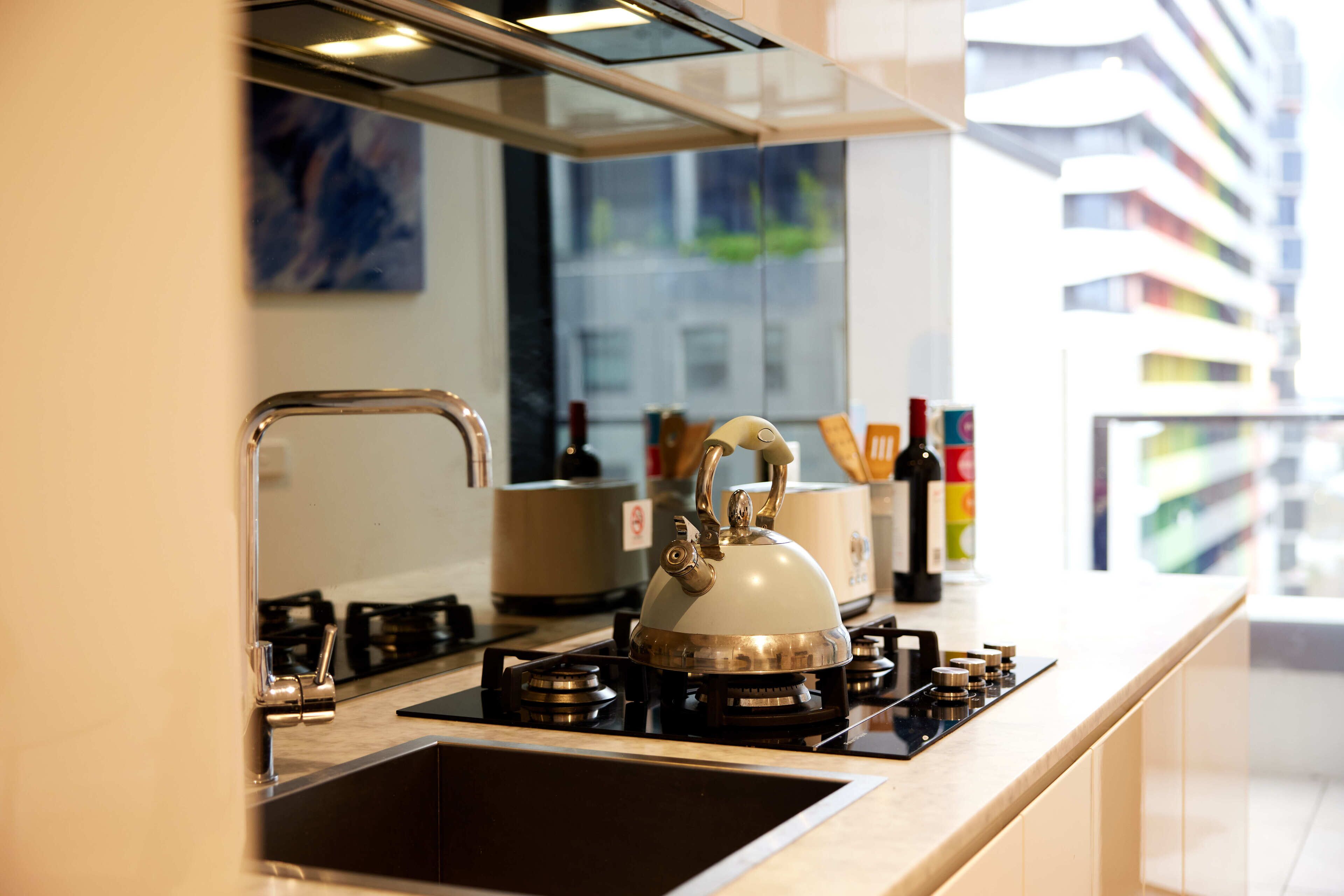 A modern kitchen with a gas stove, stainless steel kettle, and a view of a cityscape through large windows.