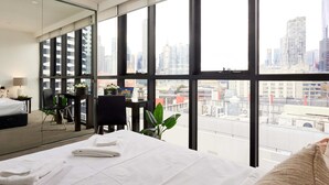 A modern hotel room with a large bed, a desk, and a view of the cityscape through large windows.