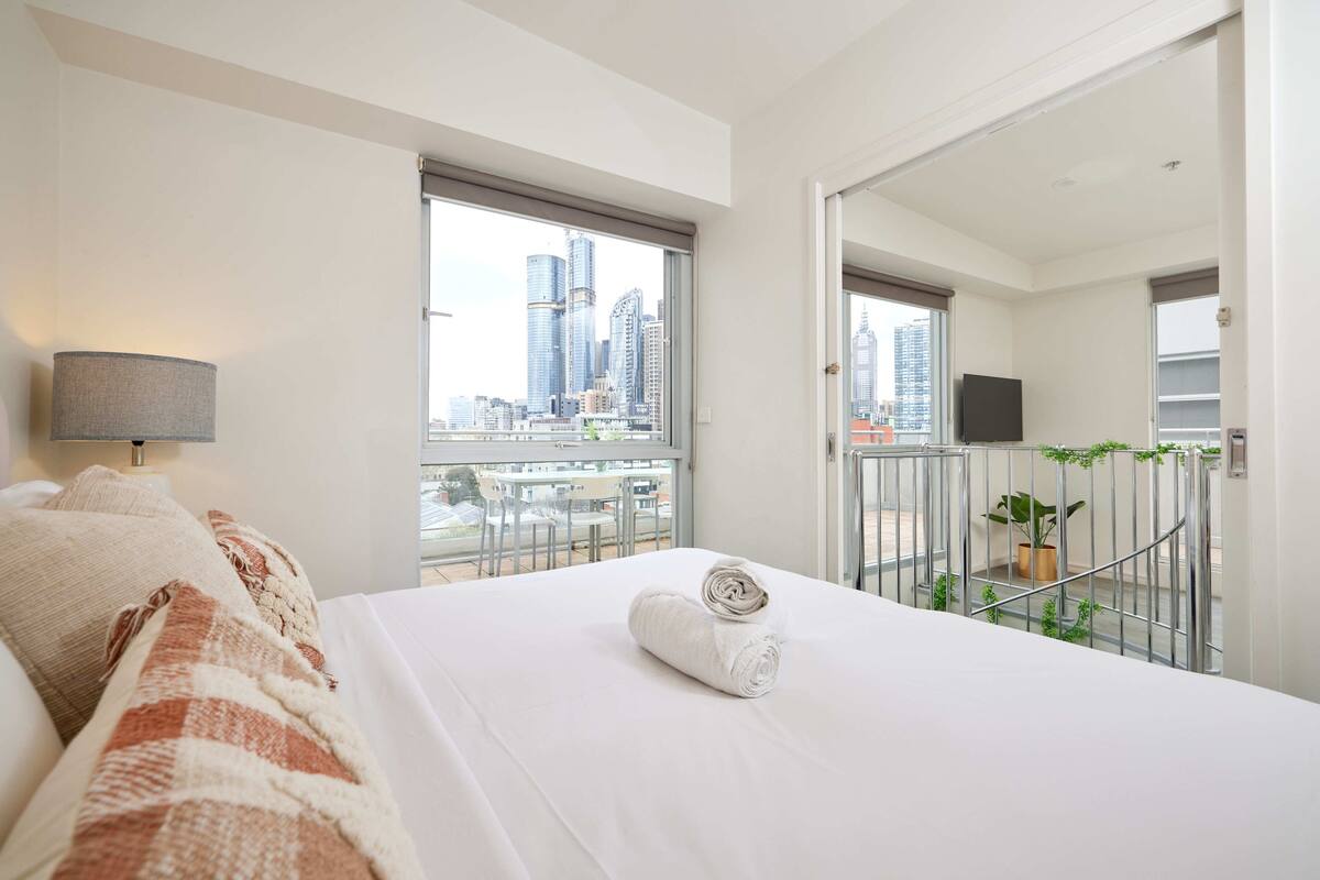 Una habitación de hotel con una cama, vistas de la ciudad, un balcón con barandilla y una planta en maceta.