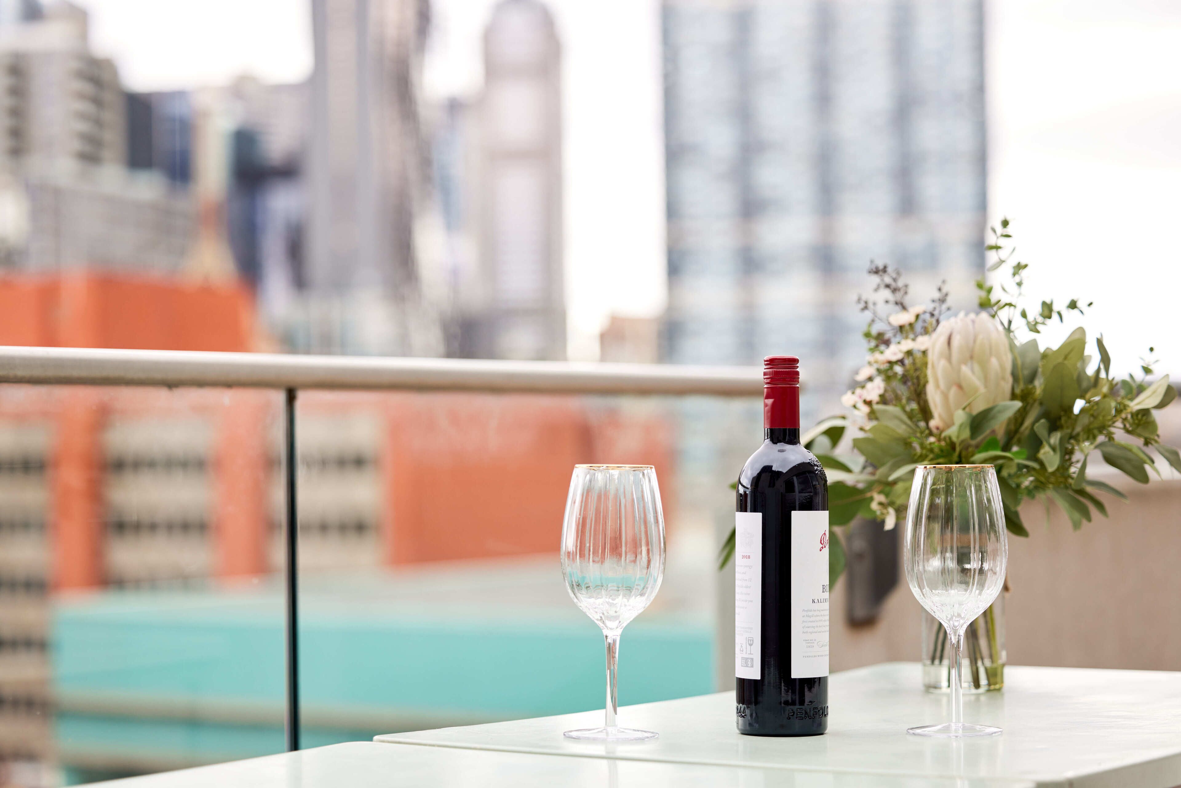 Una mesa con una botella de vino y dos copas, un florero con flores y una vista panorámica de la ciudad.