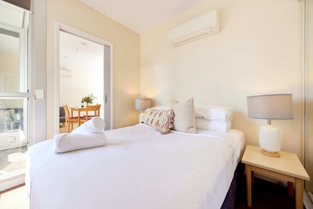 Habitación de hotel con una cama bien tendida, una mesita de noche con lámpara y vistas al exterior a través de una puerta.