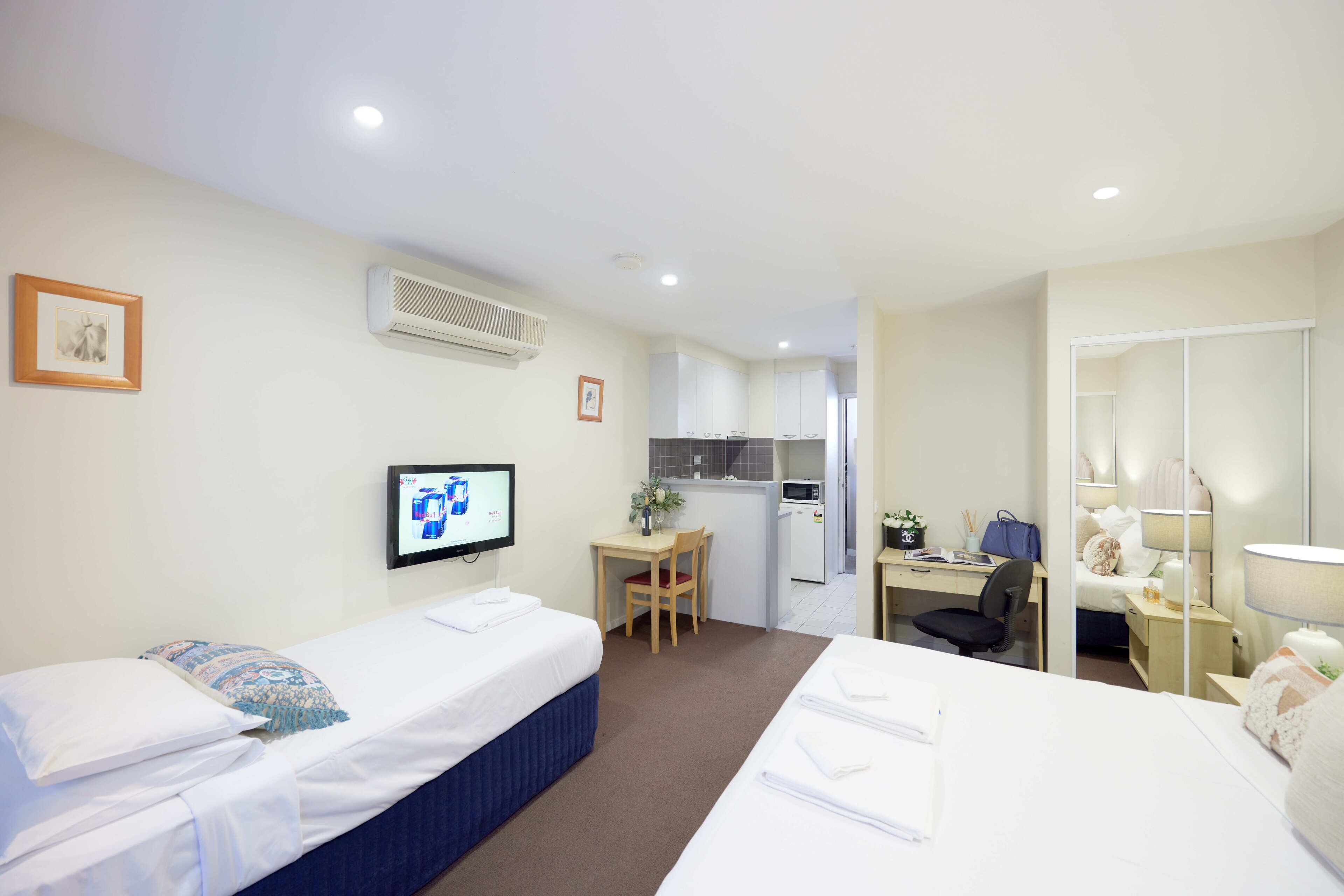 Quarto de hotel com duas camas, uma escrivaninha, uma cadeira, uma kitchenette pequena e uma televisão na parede.
