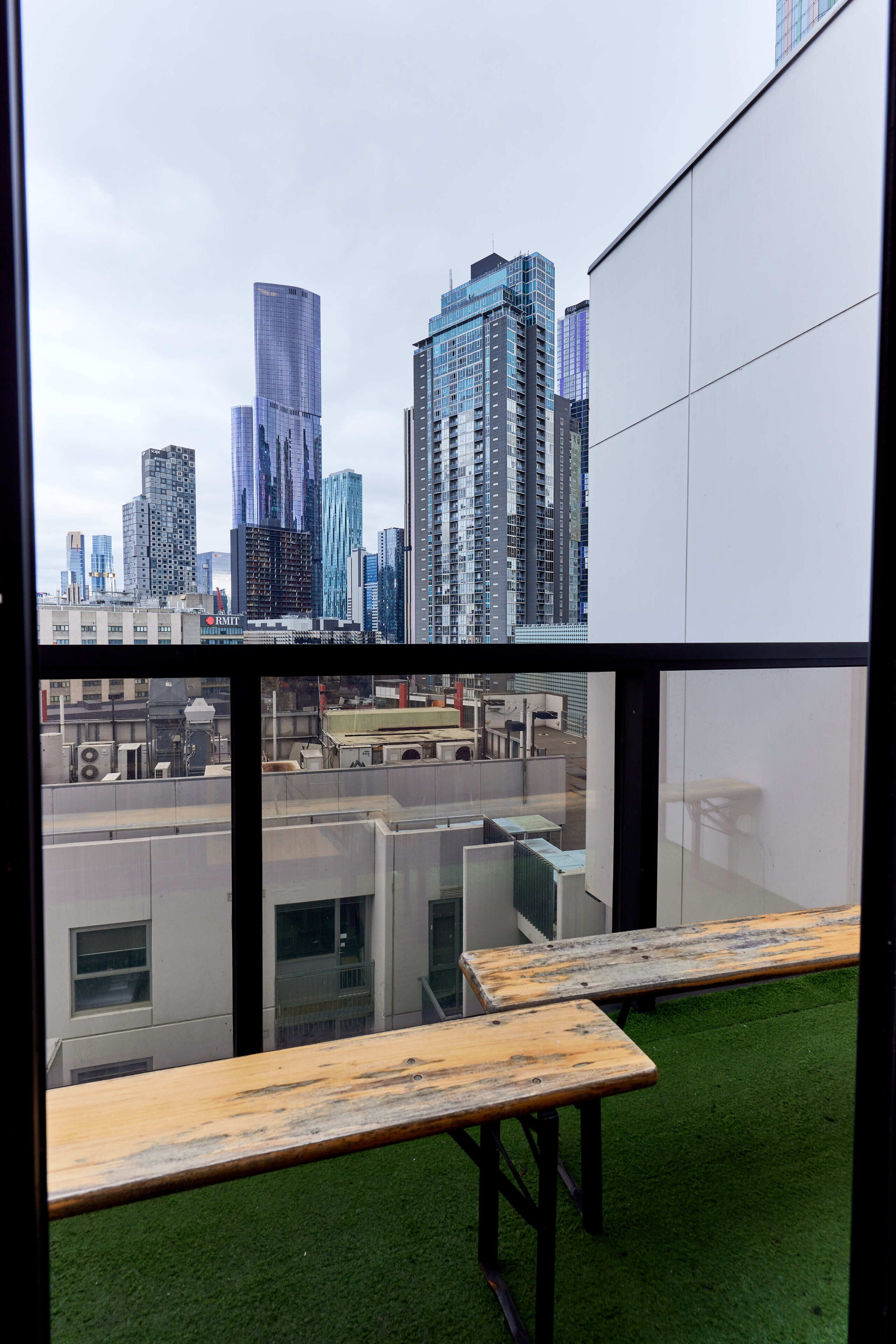 Blick auf eine Stadtlandschaft durch ein Fenster, mit modernen Hochhäusern und einer Dachterrasse mit Holztisch und Stühlen.