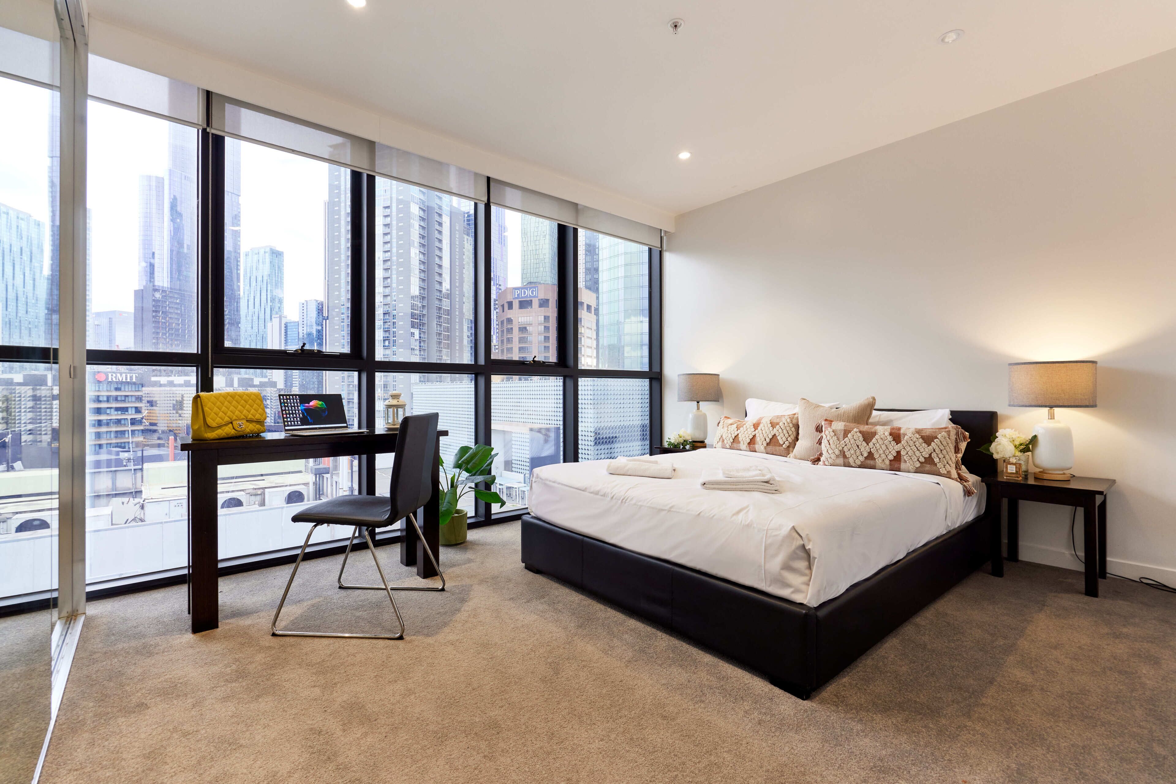 Un dormitorio moderno con una cama grande, un escritorio y vistas a un paisaje urbano a través de ventanales de piso a techo.