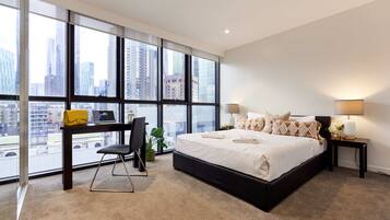 Quarto moderno com uma cama grande, uma escrivaninha e vista para a cidade através de janelas do chão ao teto.