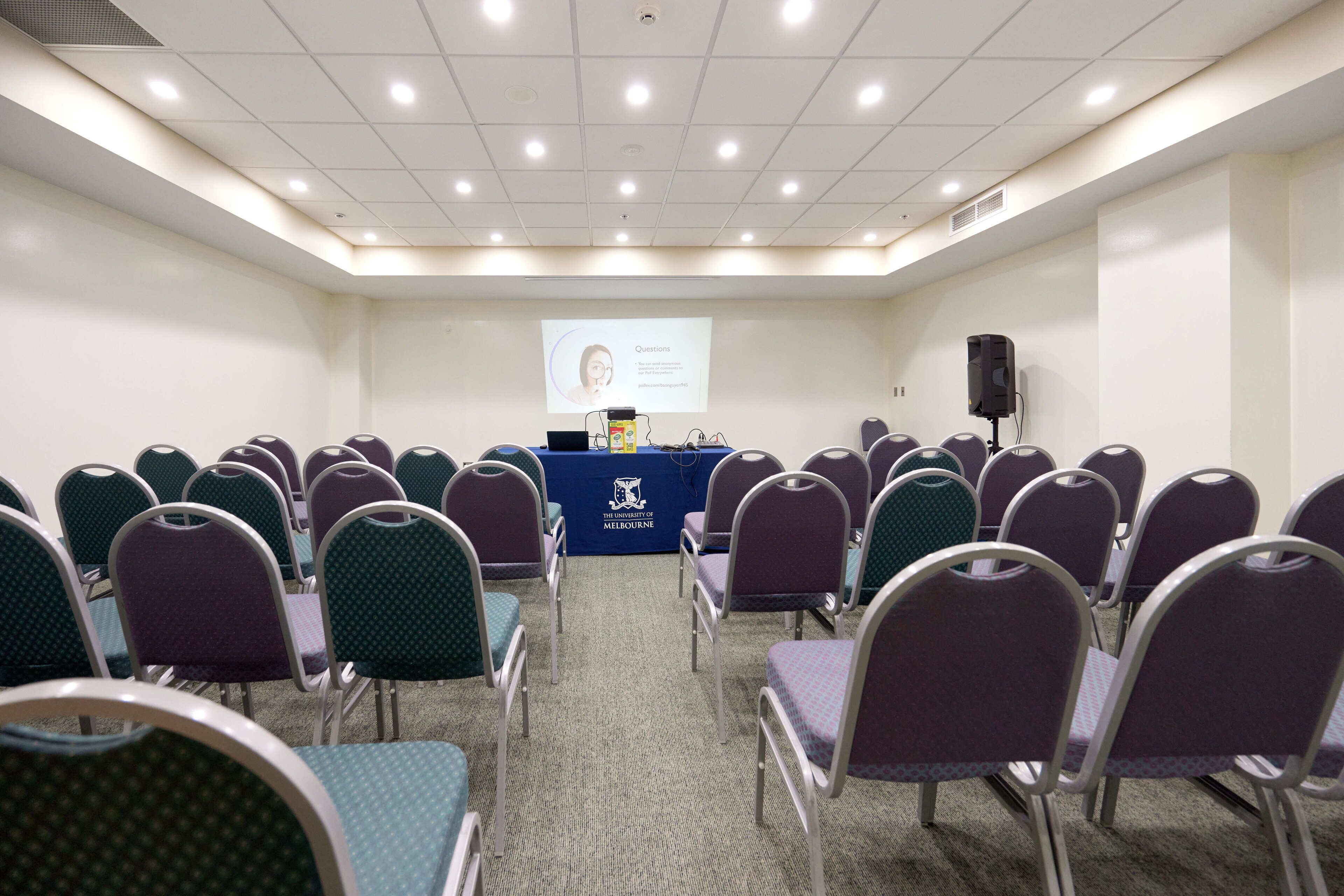 Une salle de conférence avec des rangées de chaises disposées face à un pupitre, un écran de projection et un intervenant.