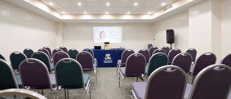 Une salle de conférence avec des rangées de chaises disposées face à un pupitre, un écran de projection et un intervenant.