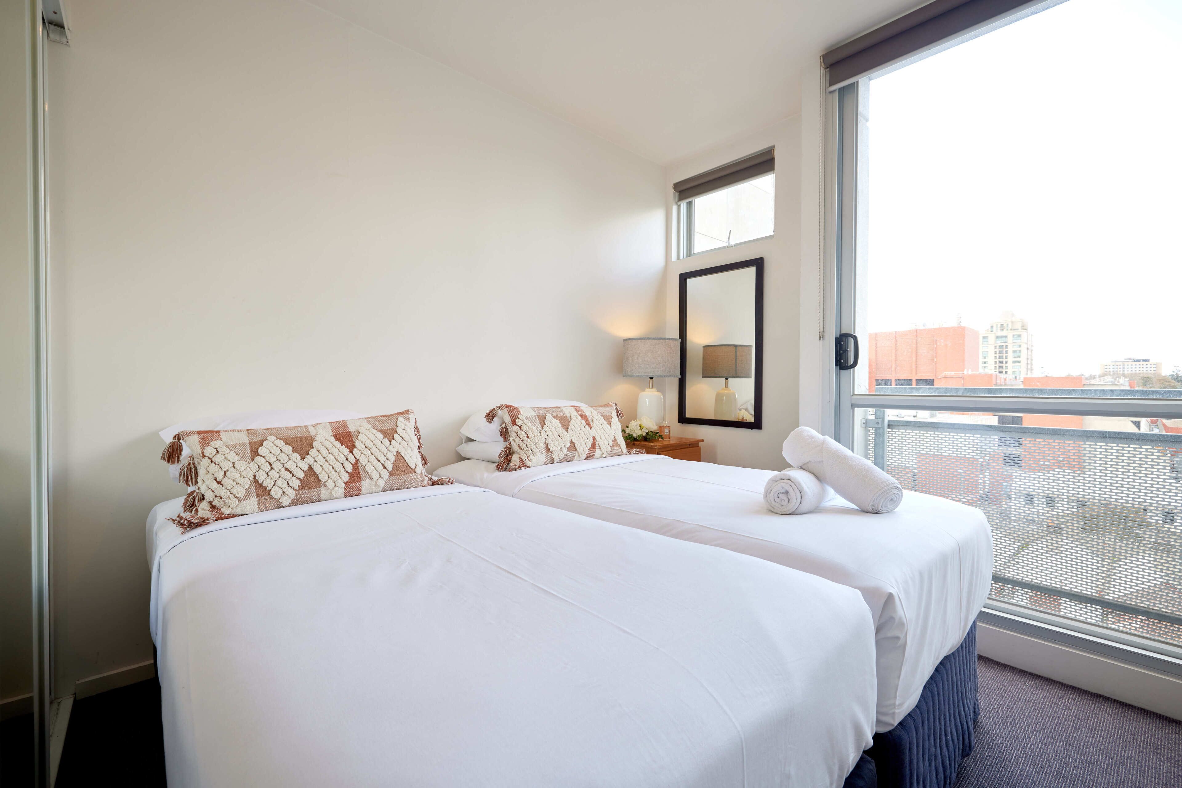A hotel room with two beds, a large window, a nightstand with a lamp, and a view of the city.