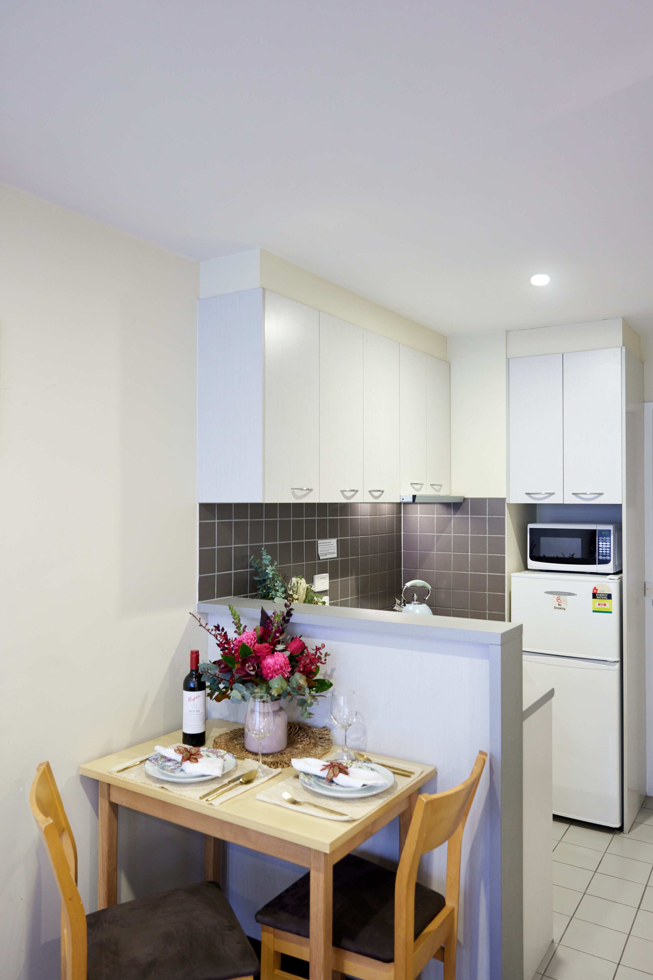 Cocina compacta con armarios blancos, microondas y un refrigerador pequeño. Una mesa de comedor con sillas y un florero están colocados en el centro.