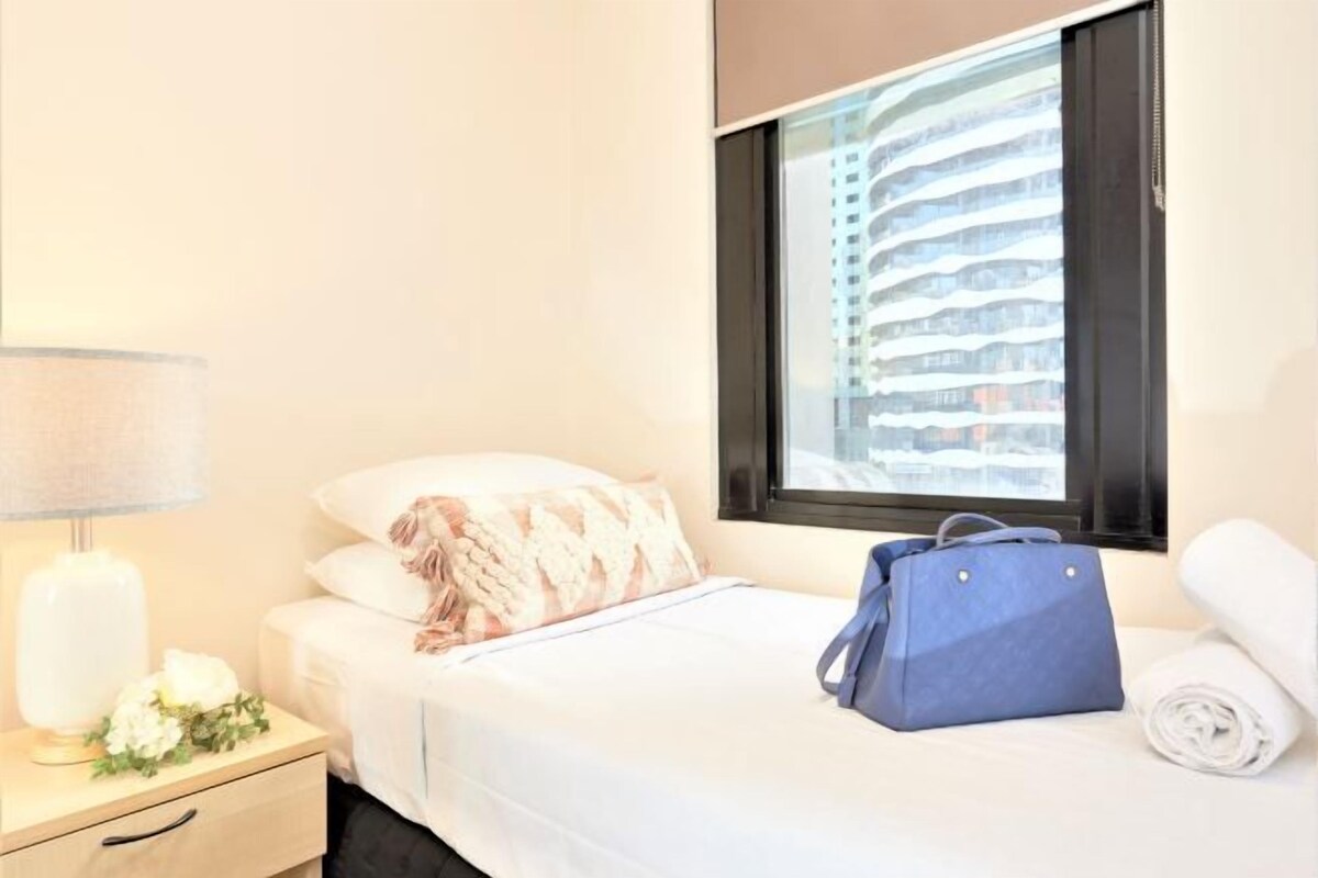 A hotel room with a bed, a nightstand with a lamp, a window with a view of buildings, and a blue bag on the bed.