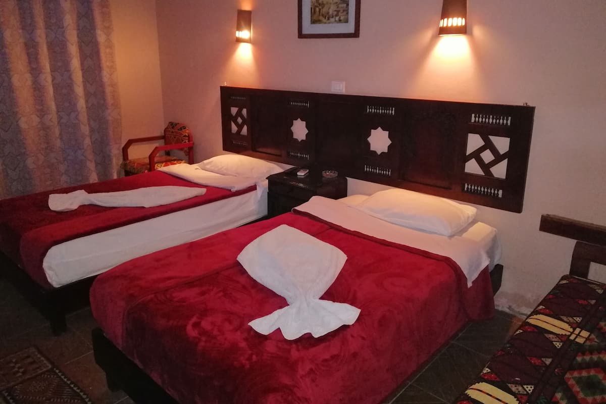 Quarto de hotel com duas camas, cada uma com colcha vermelha e almofadas brancas. Há duas luzes embutidas na parede e um quadro emoldurado acima das camas.
