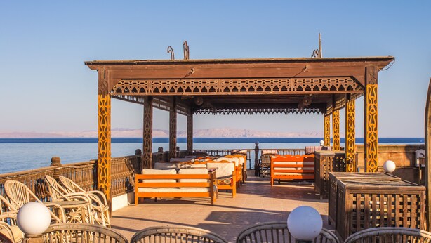 A wooden gazebo with benches and wicker chairs overlooking a body of water with mountains in the background. - Ali Baba Hotel (Dahab)